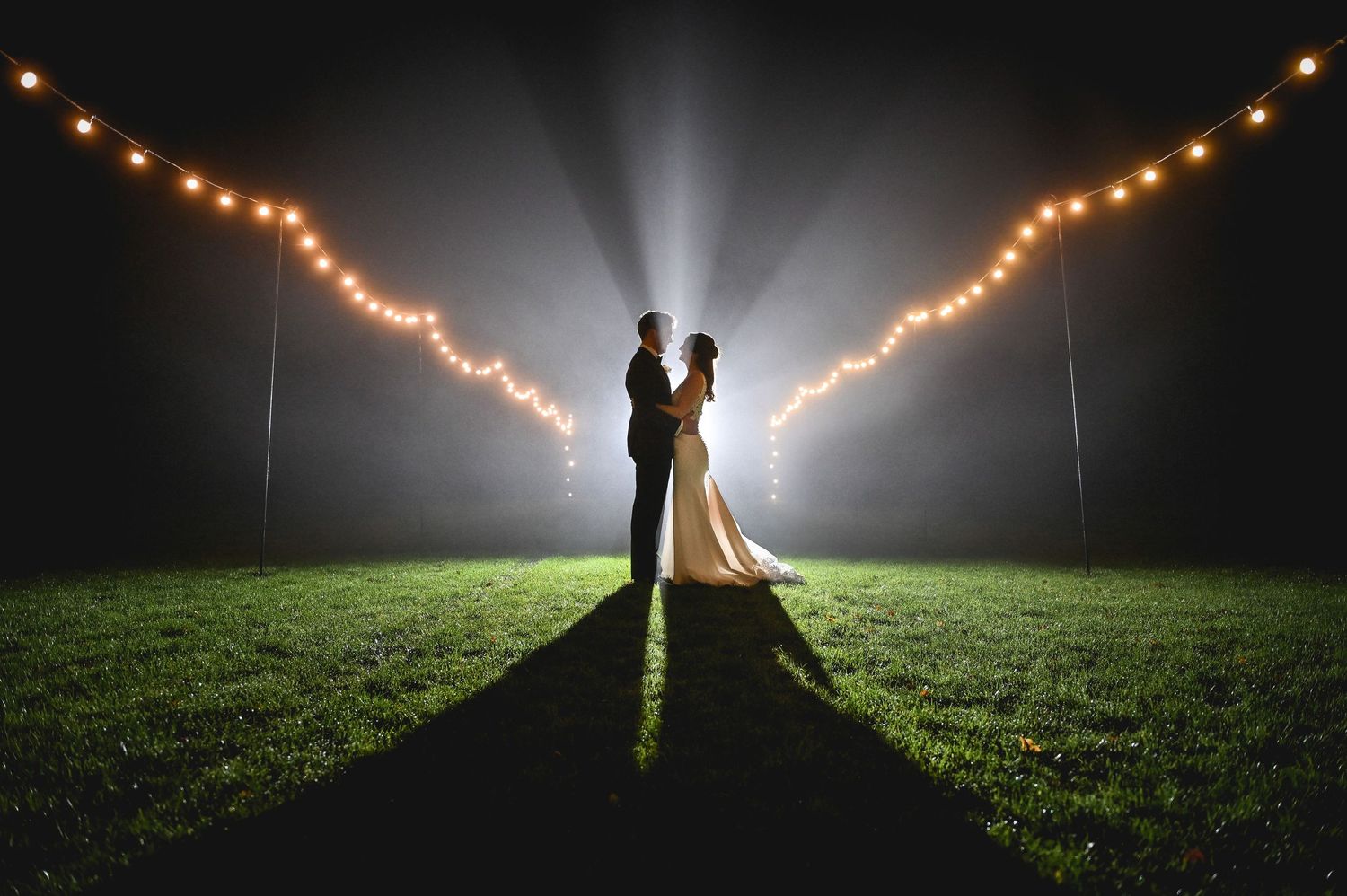 A romantic nighttime wedding photo with string lights creating a V-shape behind the couple on a grassy field.