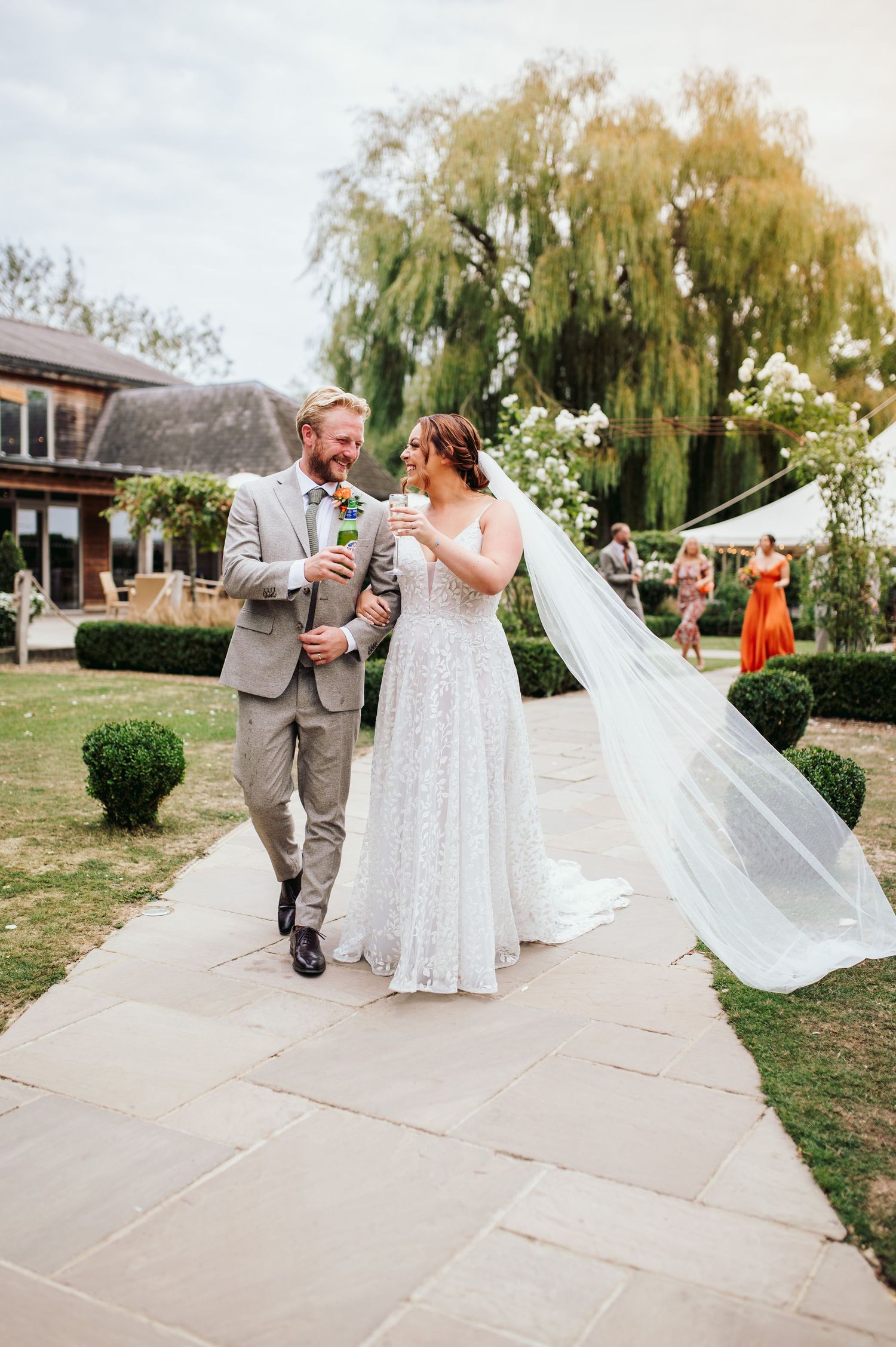 A couple walks along a stone pathway with willow trees in the background during their wedding celebration.