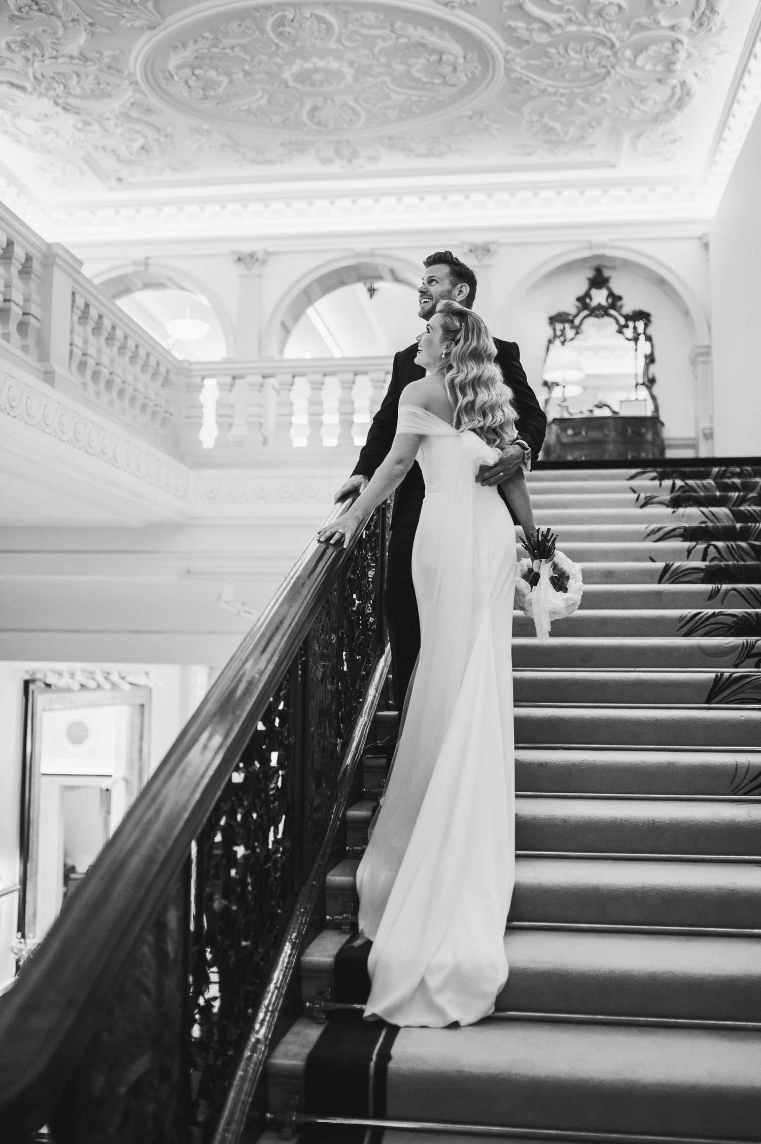 Elegant black and white photo of a couple on a grand staircase with ornate ceiling and architecture.