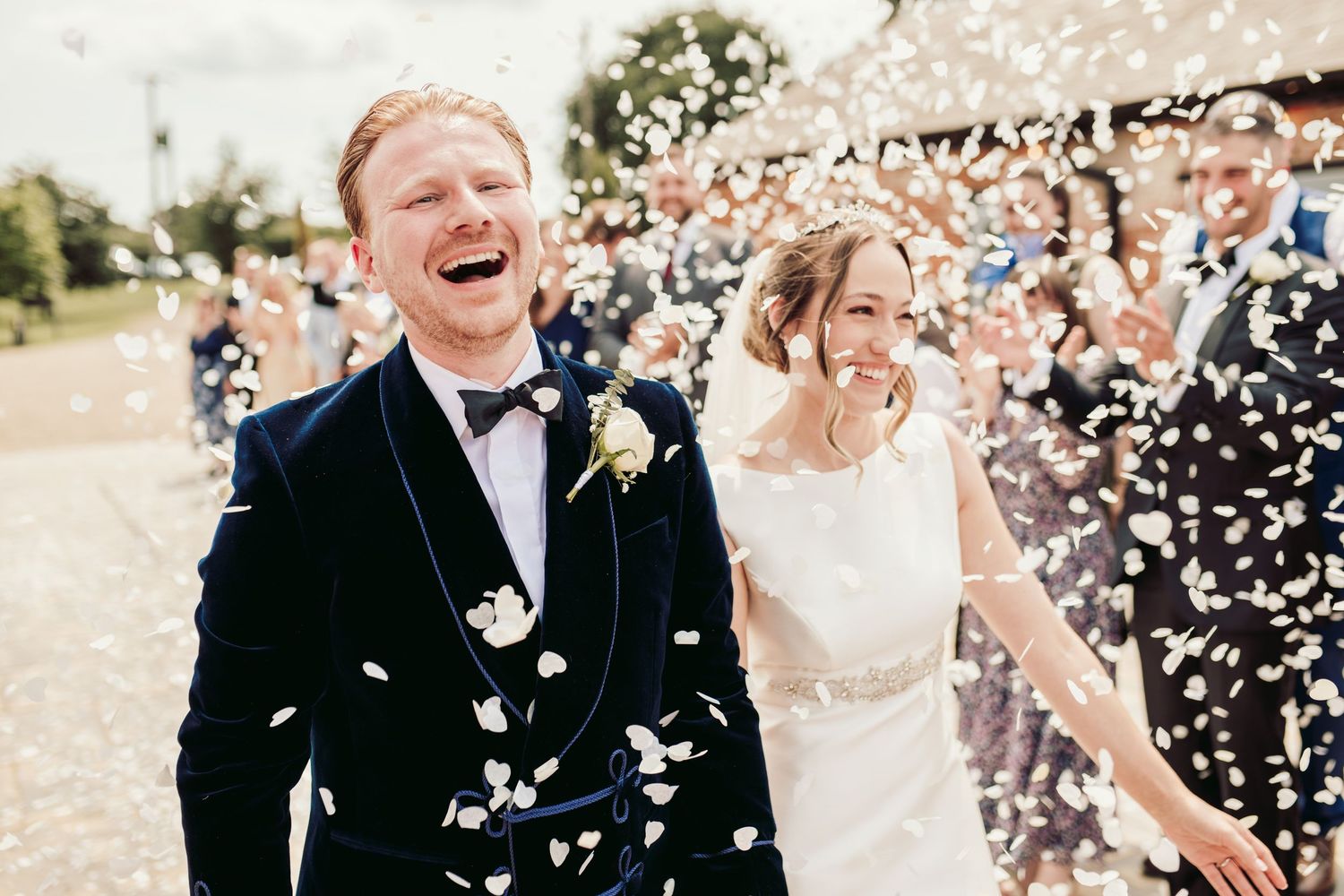 Wedding celebration with white flower petals being tossed in the air as newlyweds share joyful moments outdoors.