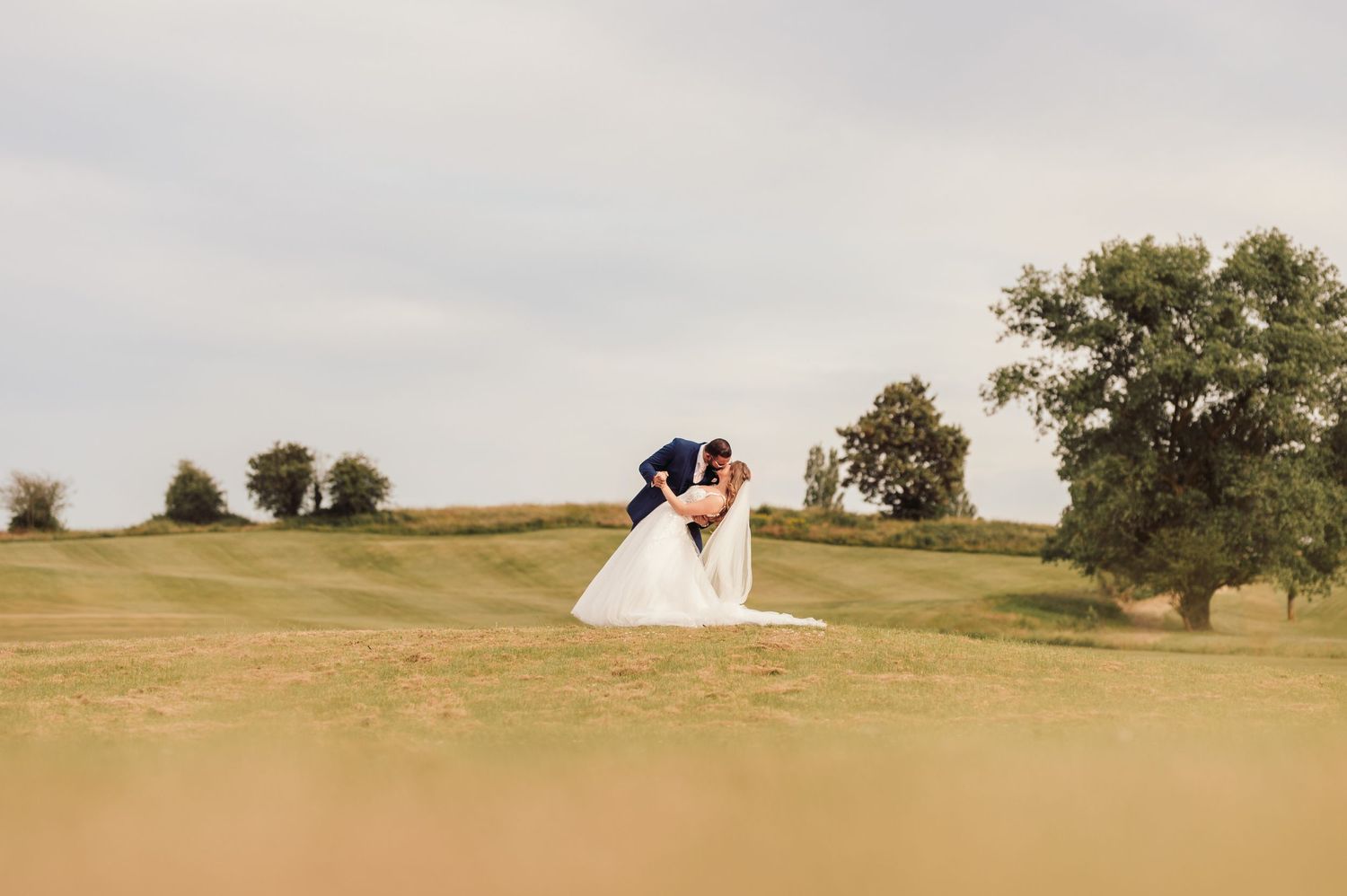 A couple embraces in a romantic moment on rolling hills of a golf course with trees in the background at golden hour.