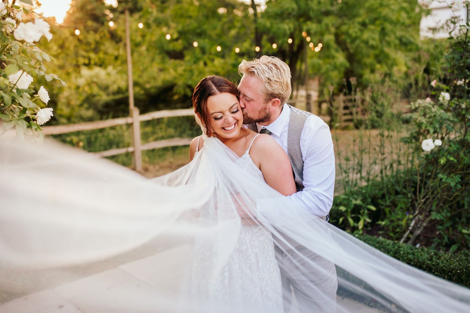 A romantic wedding couple embraces while a flowing veil catches the breeze in an outdoor setting.