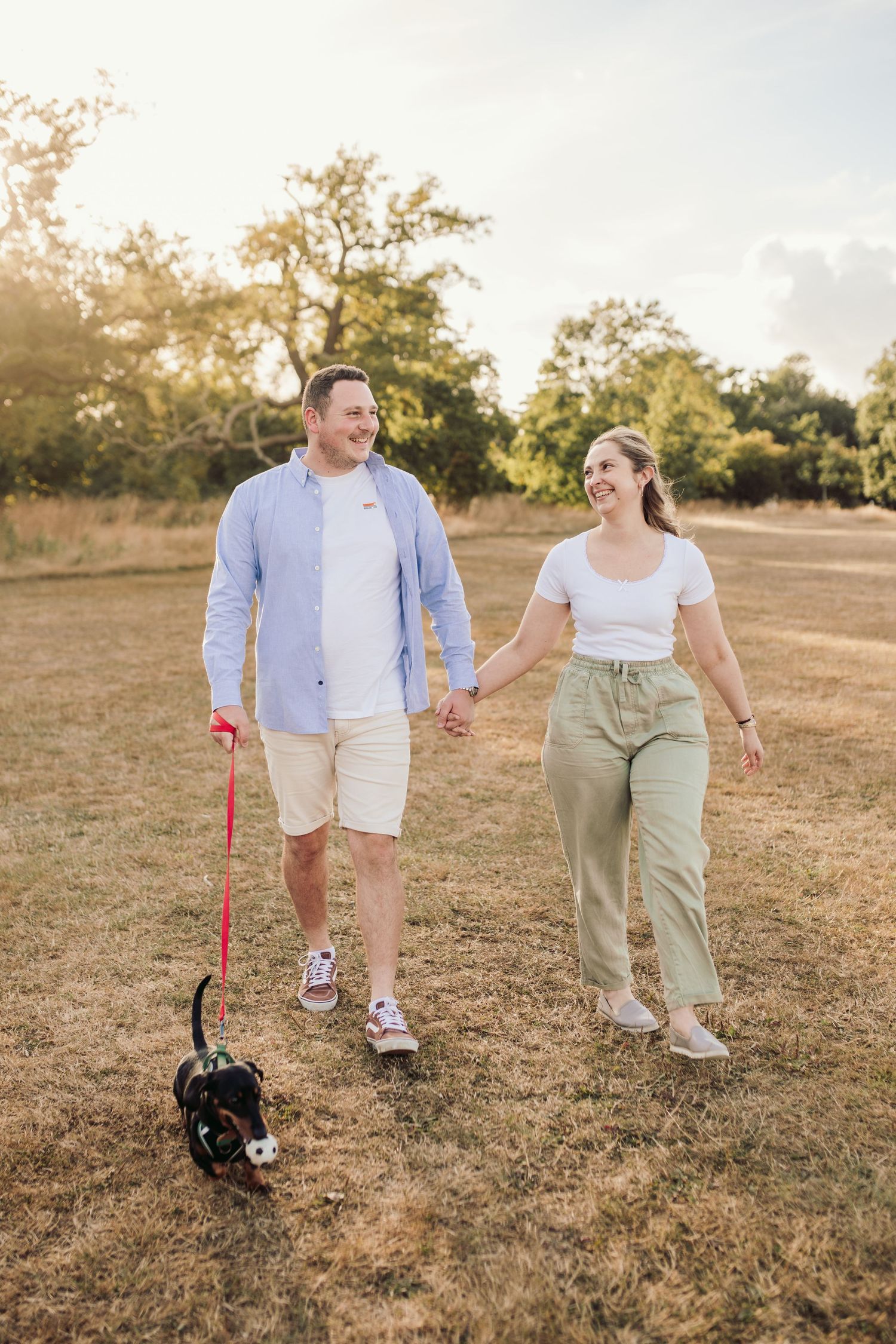 A couple walks their small black dog on a sunny trail through a grassy field.