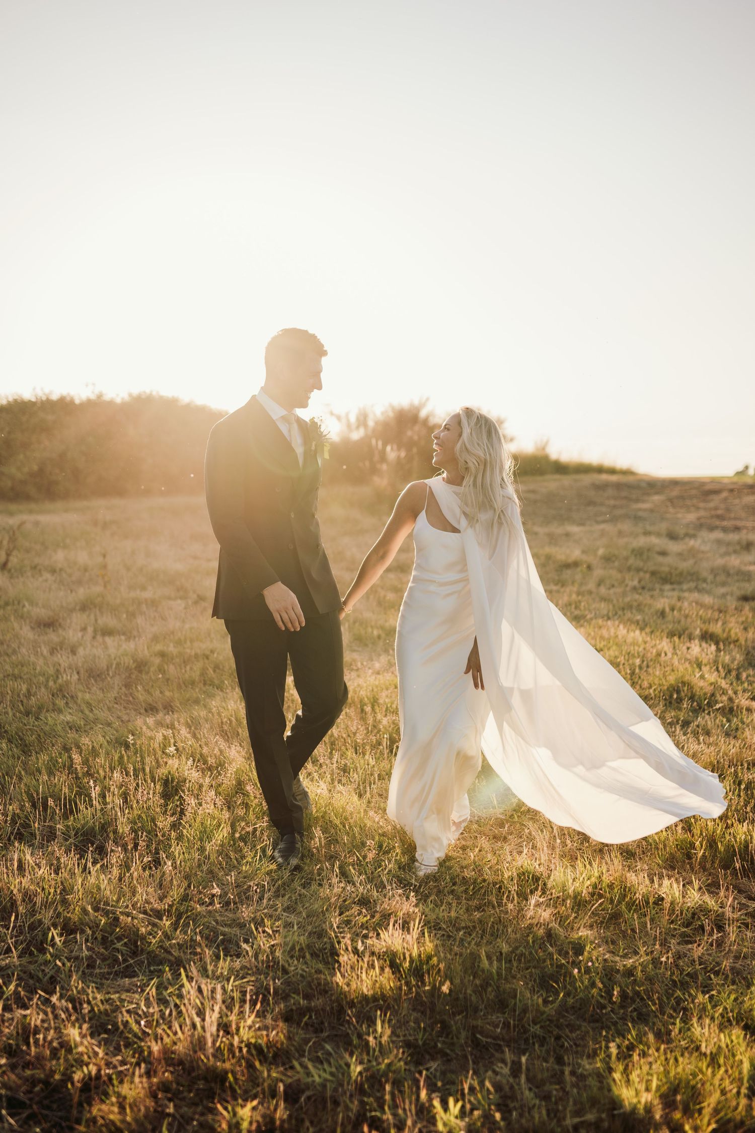 Silhouetted couple walks through sunlit field in wedding attire.