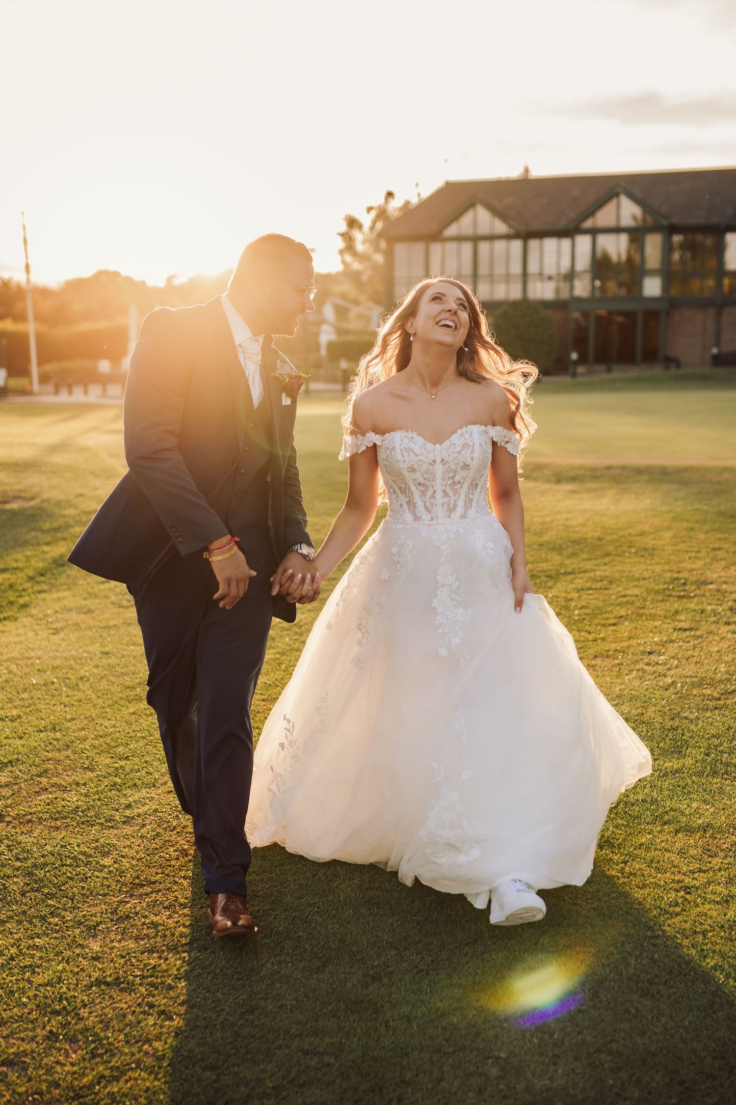 A couple shares a joyful moment walking on a golf course during golden hour sunset, bride wearing an off-shoulder lace gown.