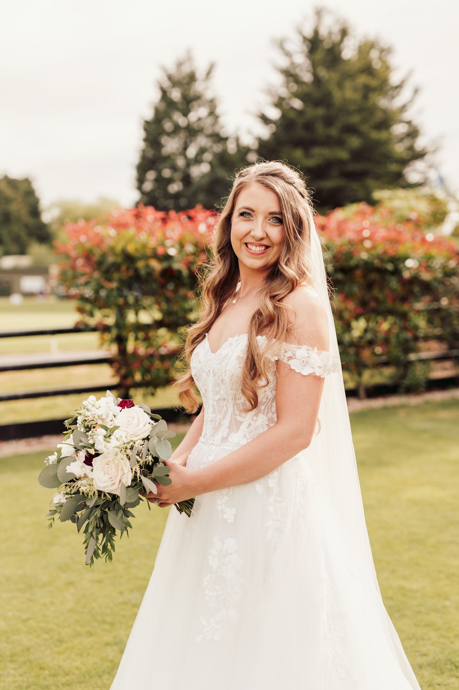 A bride in an off-shoulder lace wedding dress holds a white floral bouquet on a lawn with blooming flowers and trees.