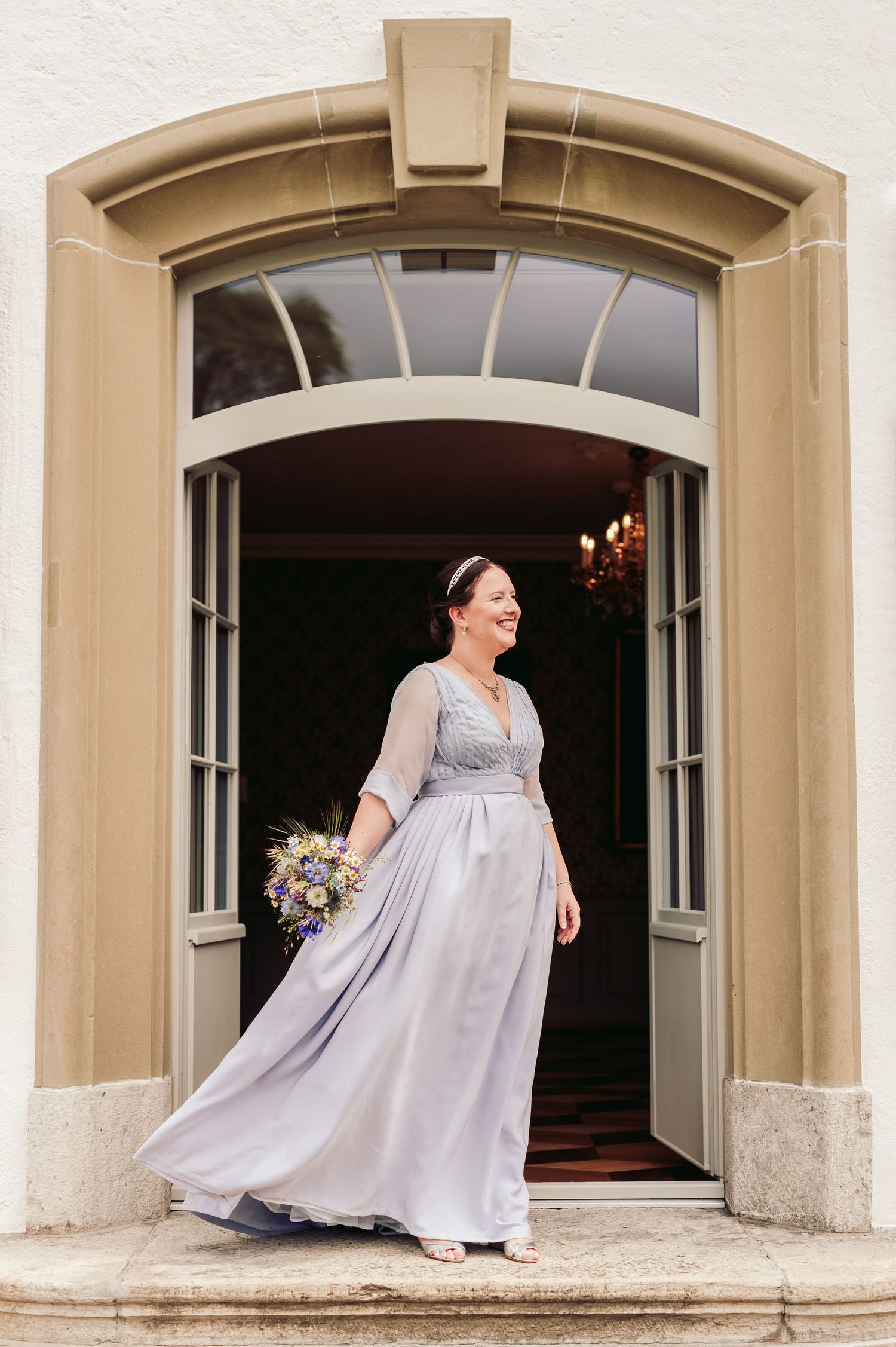 A person in an elegant flowing gray dress and bouquet stands in an arched beige doorway entrance. Spiez switzerland destination wedding