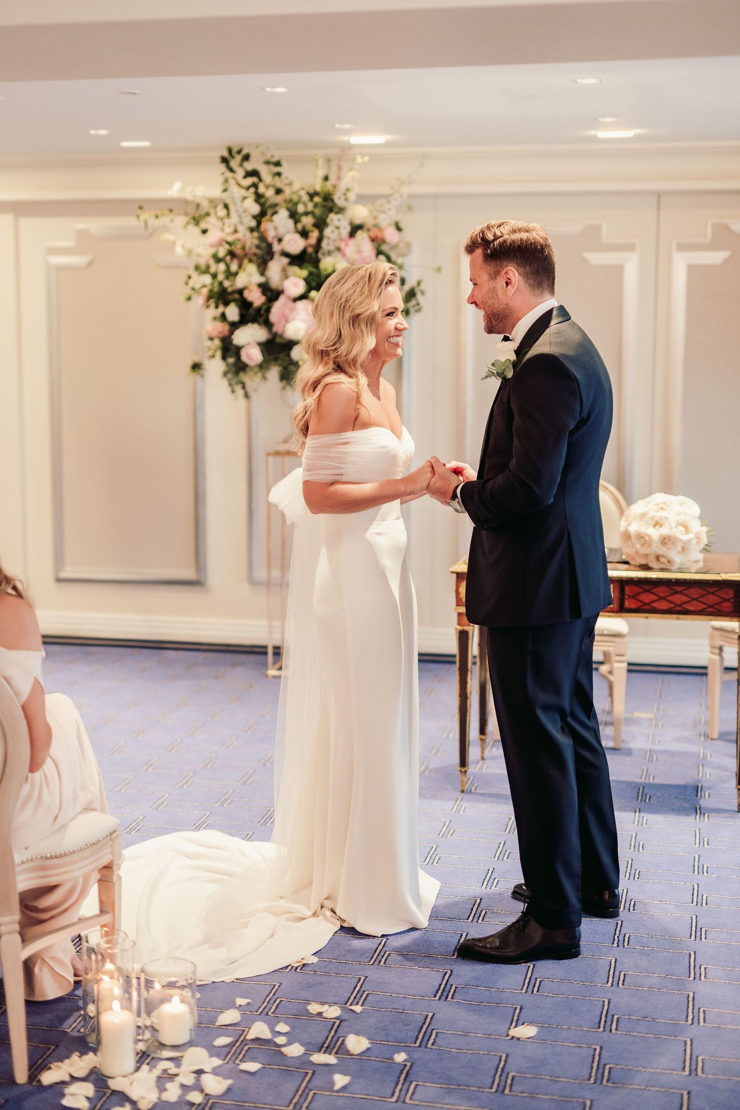 A couple shares a tender moment during their wedding ceremony in an elegant room with floral arrangements and blue carpeting.Claridge’s