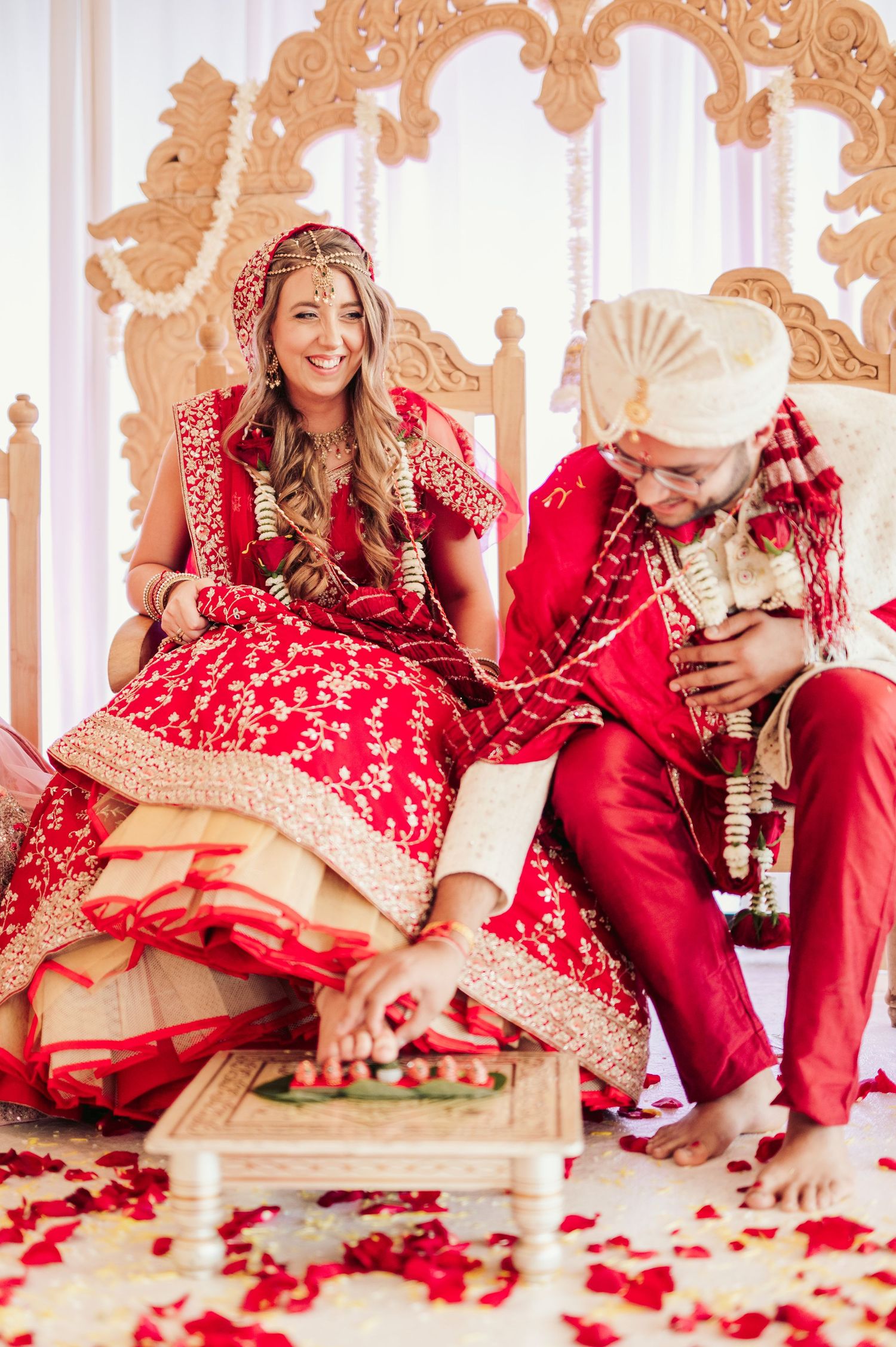 Traditional Indian wedding ceremony on decorative throne with couple in red attire surrounded by rose petals.