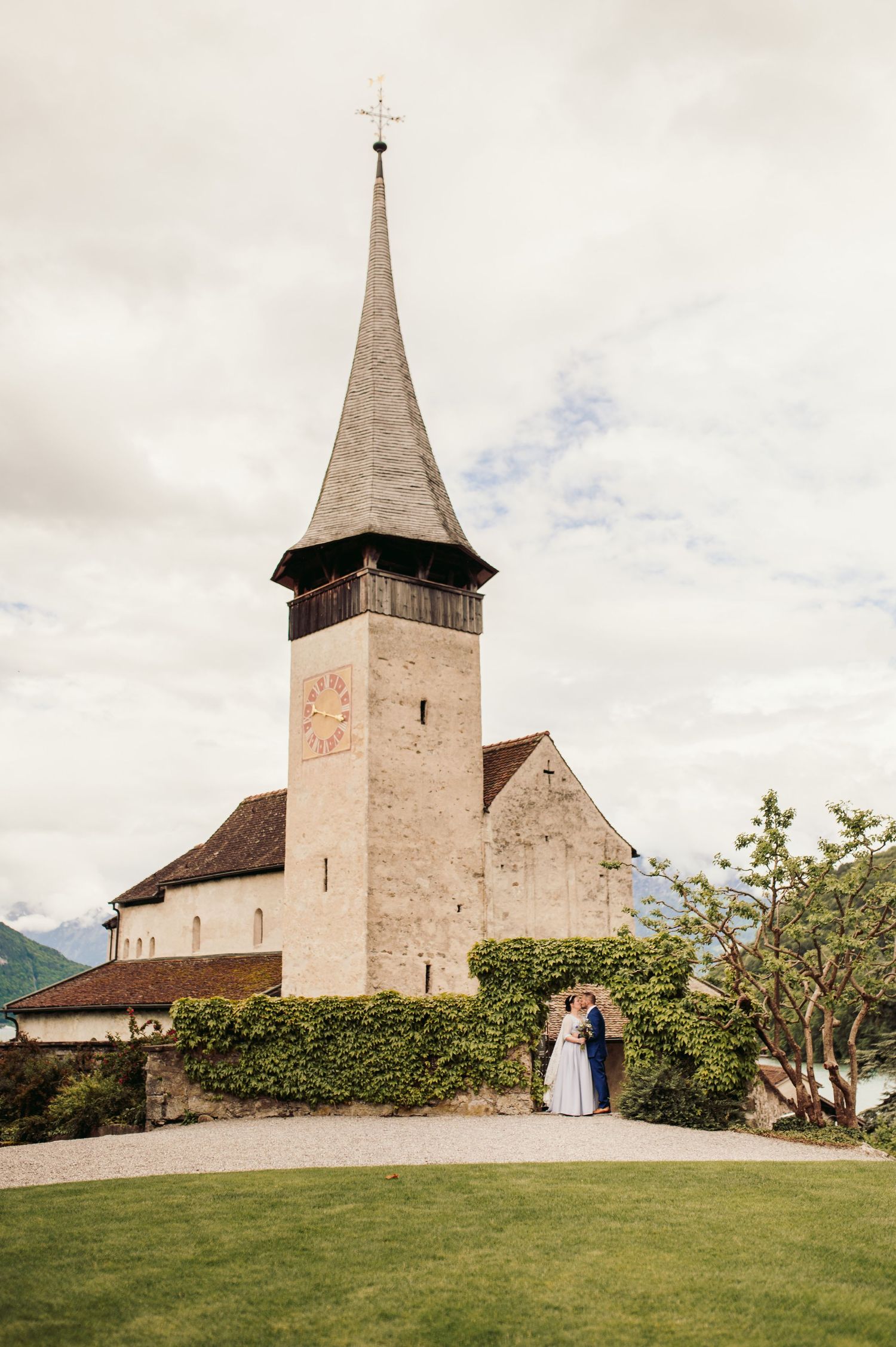 A picturesque historic church with a tall spire and clock tower stands against a cloudy sky in a scenic mountain setting. Spiez switzerland