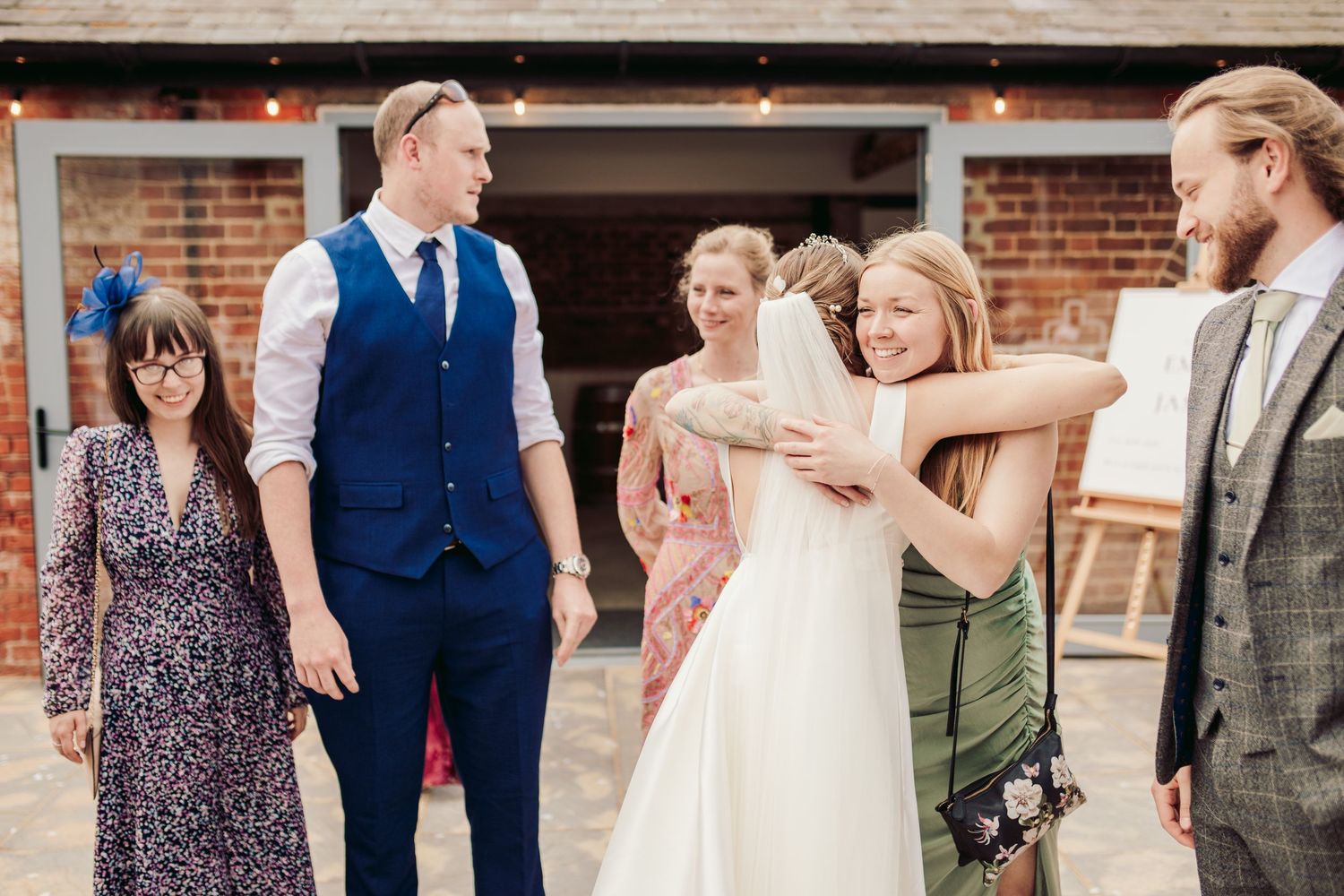 A joyful wedding celebration scene with guests gathered outside a brick building during a summer afternoon.