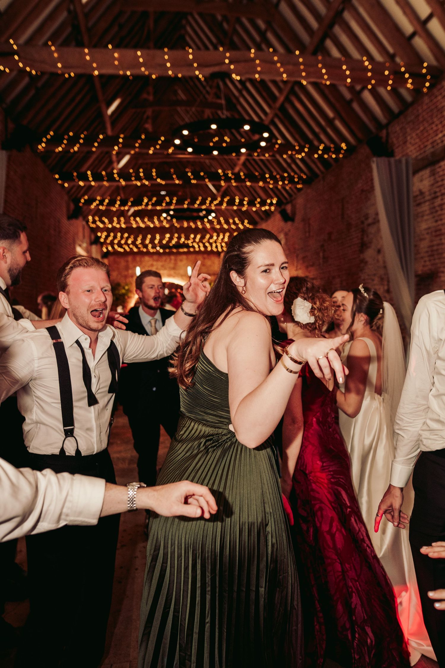 Party guests dance enthusiastically at a wedding reception under string lights in a rustic barn venue.