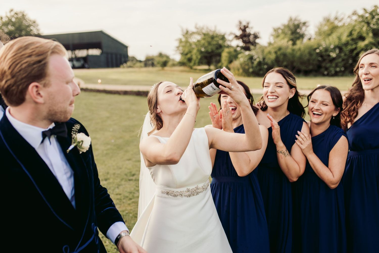 Wedding party laughs together outdoors on a sunny day wearing navy blue and white attire.