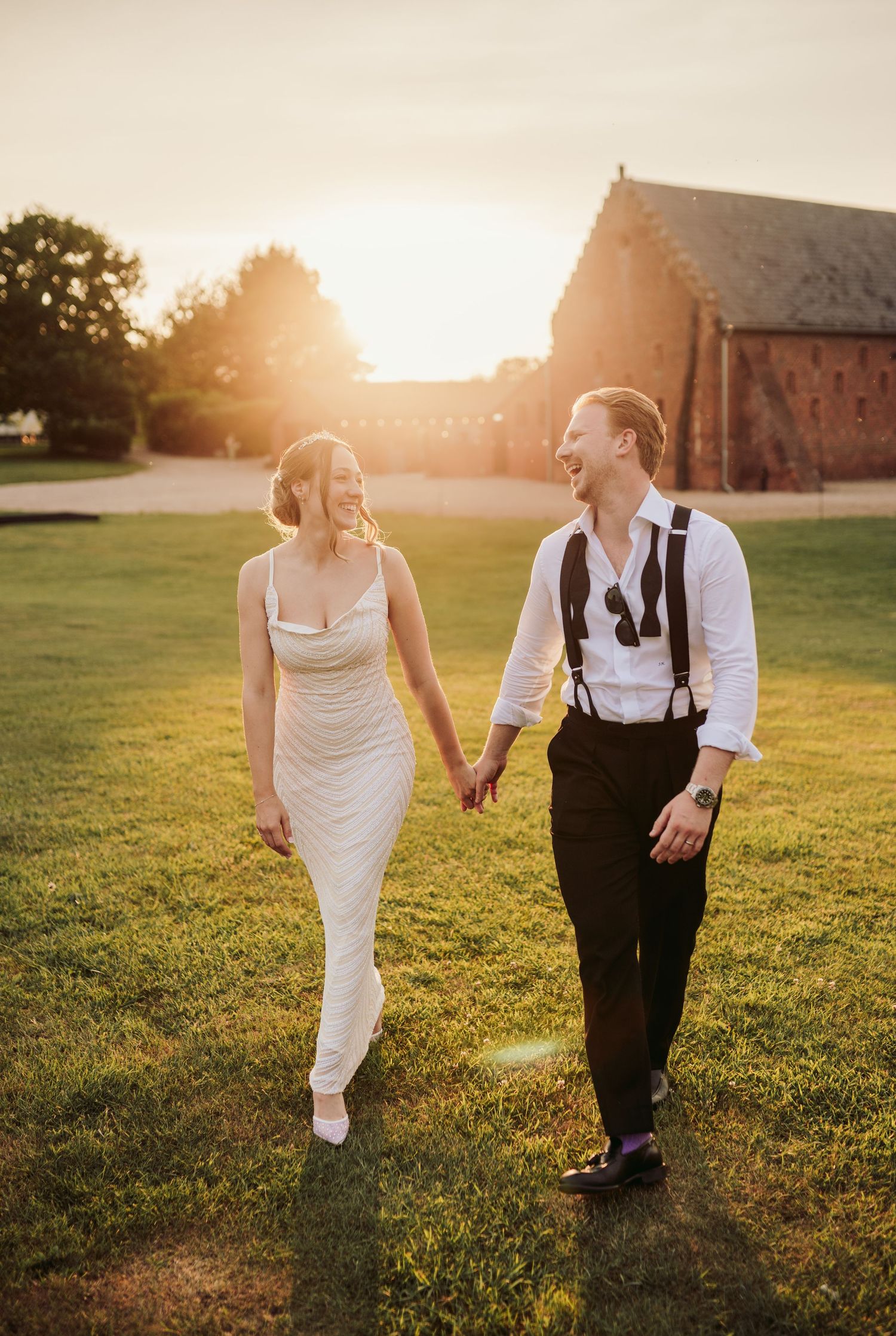 A couple walks hand in hand through a sunlit field at golden hour with a rustic barn in the background.