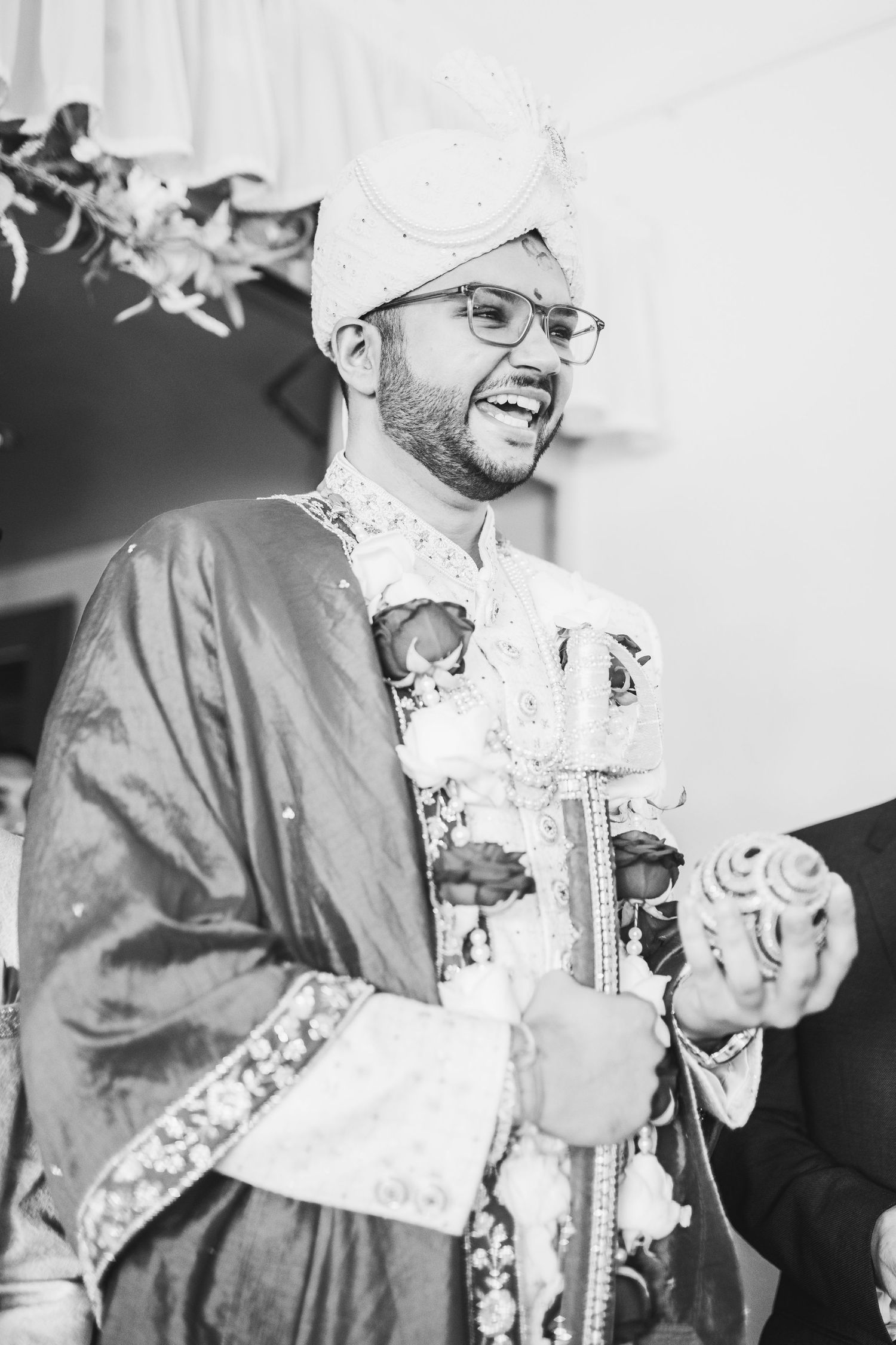Black and white portrait of a smiling person in traditional South Asian wedding attire with glasses and a white turban.