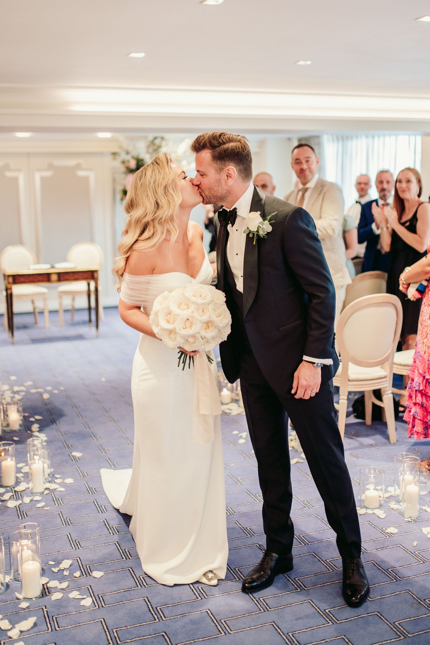 Couple shares romantic kiss during wedding ceremony in elegant ballroom with candlelit decor and guests watching.Claridge’s wedding