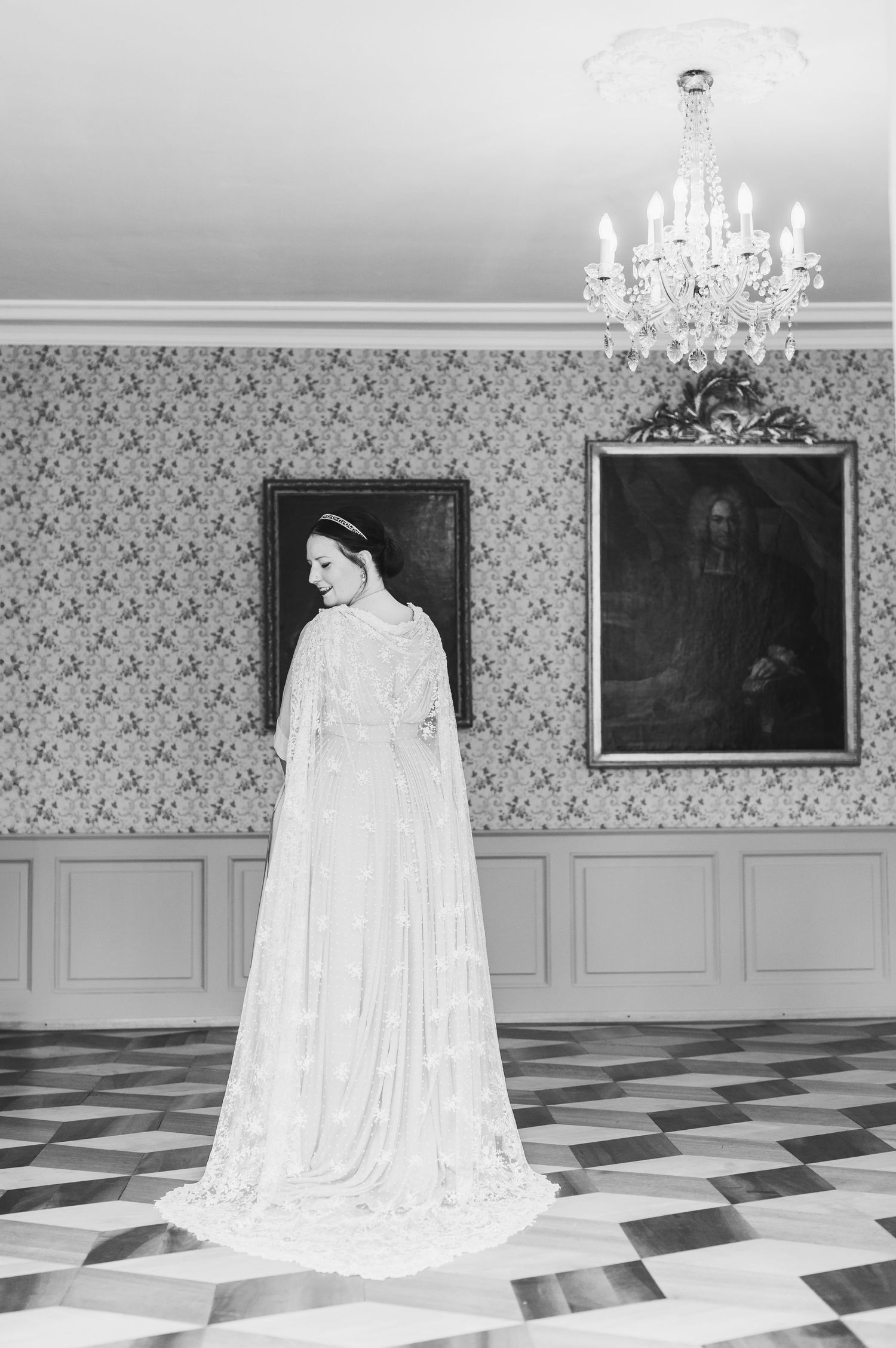 A dramatic wedding dress with a long flowing cape stands in an elegant room with checkered floors and a crystal chandelier. Spiez