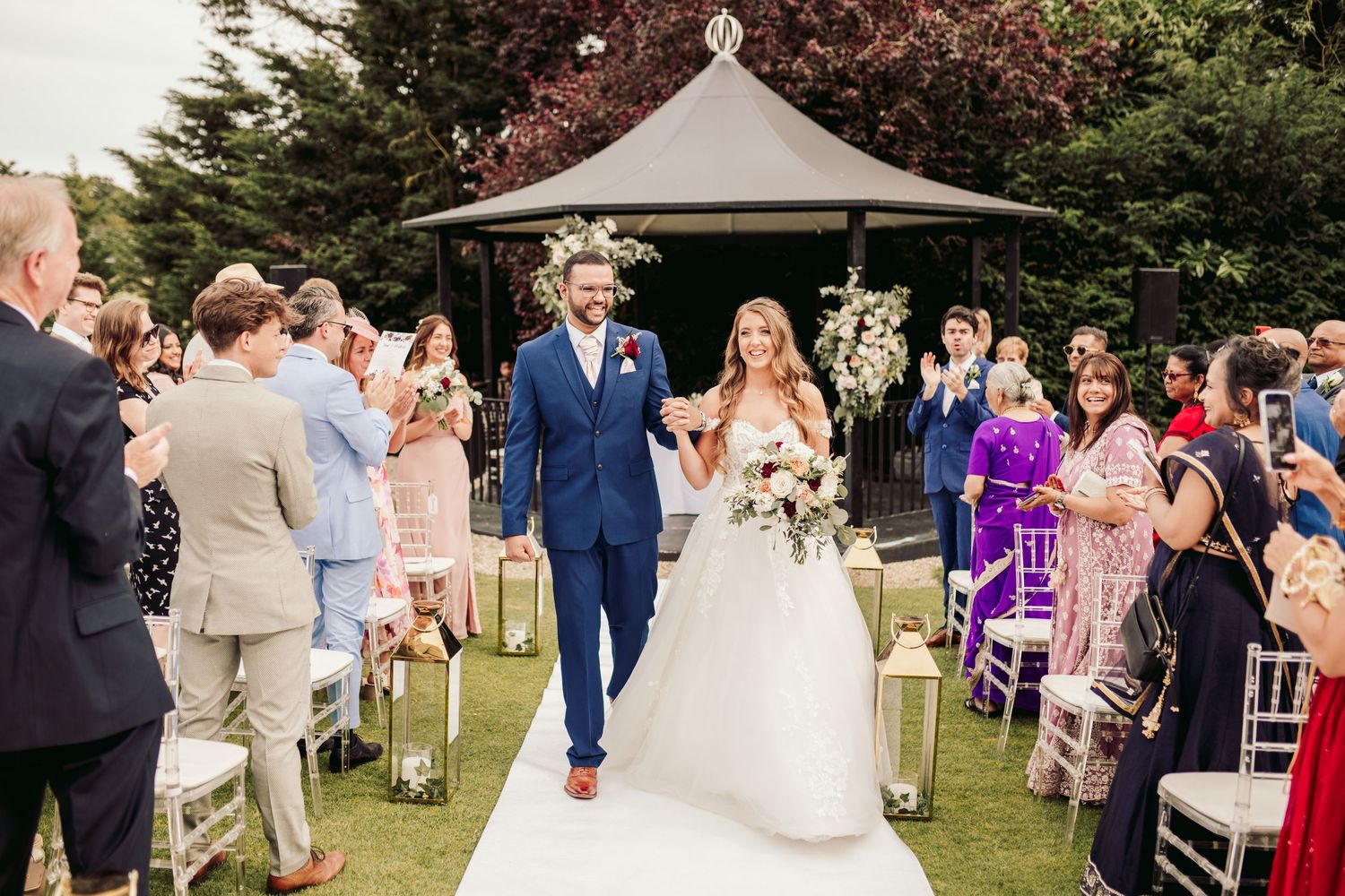 Outdoor wedding ceremony in garden setting with couple walking down white aisle runner under wooden gazebo structure.