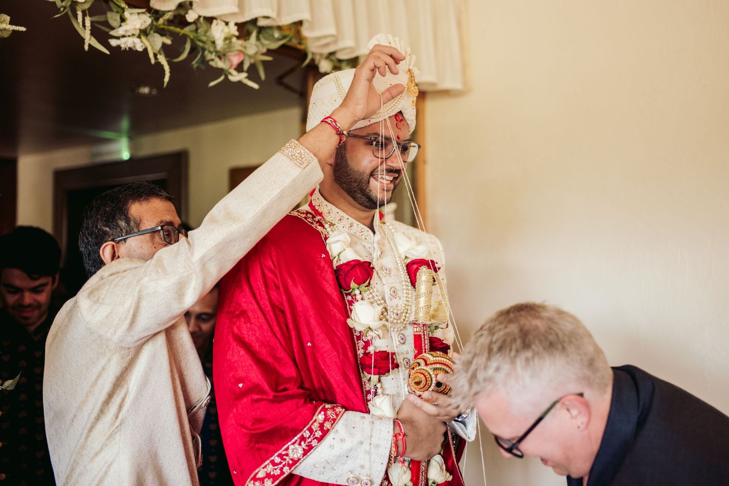Traditional Indian wedding ceremony with groom in vibrant red sherwani and floral garlands during a ritual moment.