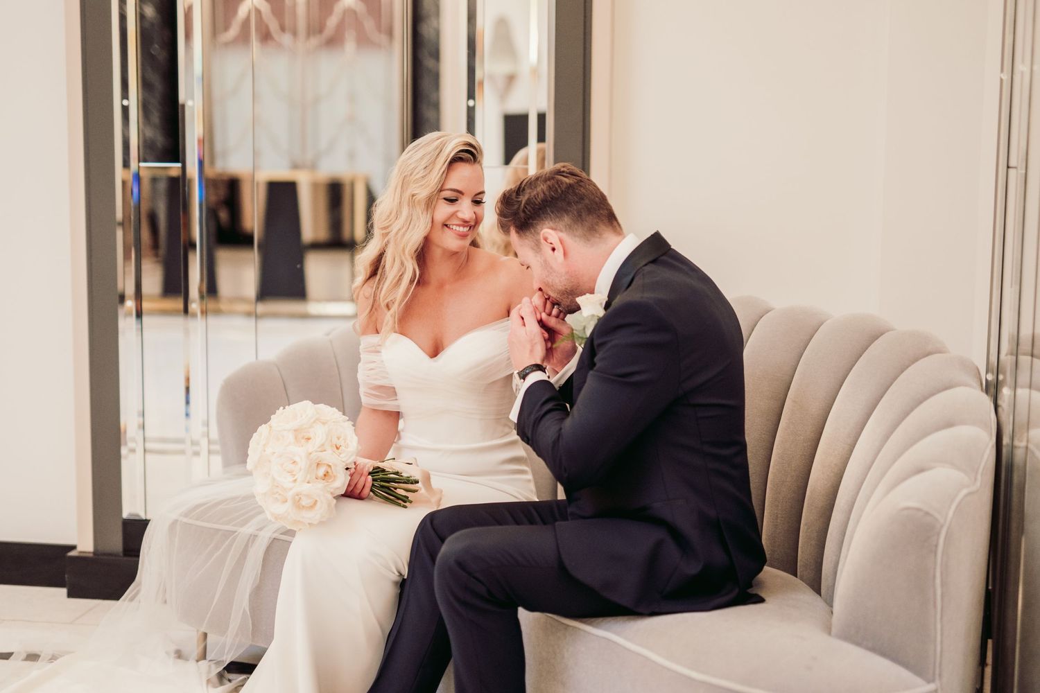 Elegant couple shares intimate moment on luxurious beige sofa with white floral bouquet during wedding celebration.Claridge’s wedding