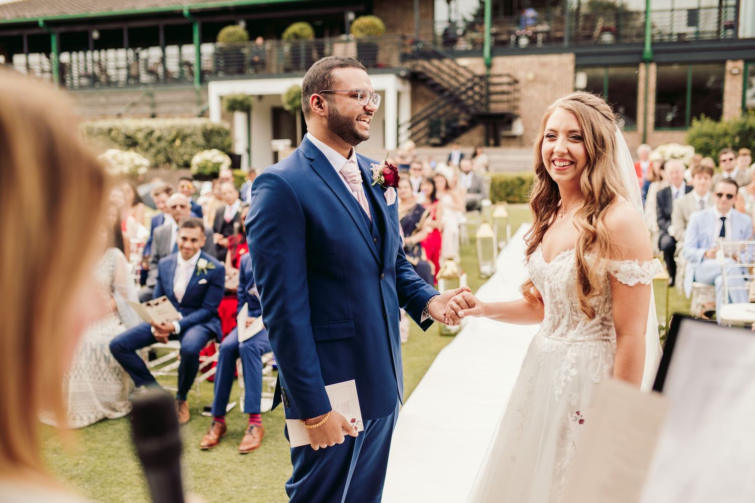 A happy couple celebrates their outdoor wedding ceremony with guests seated on the lawn below a rustic venue.