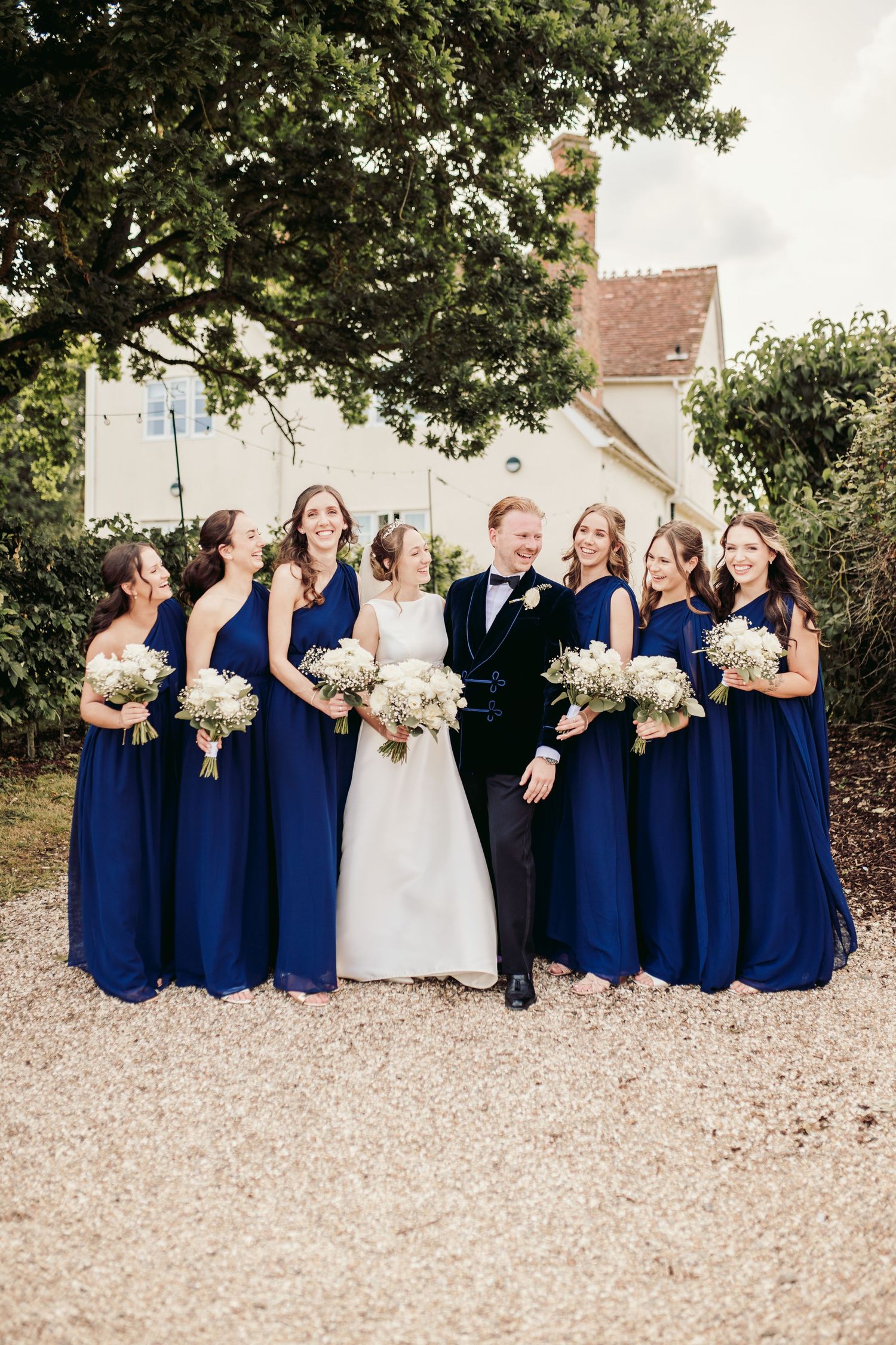 Wedding party wearing navy blue dresses and holding white bouquets pose together on a gravel path.