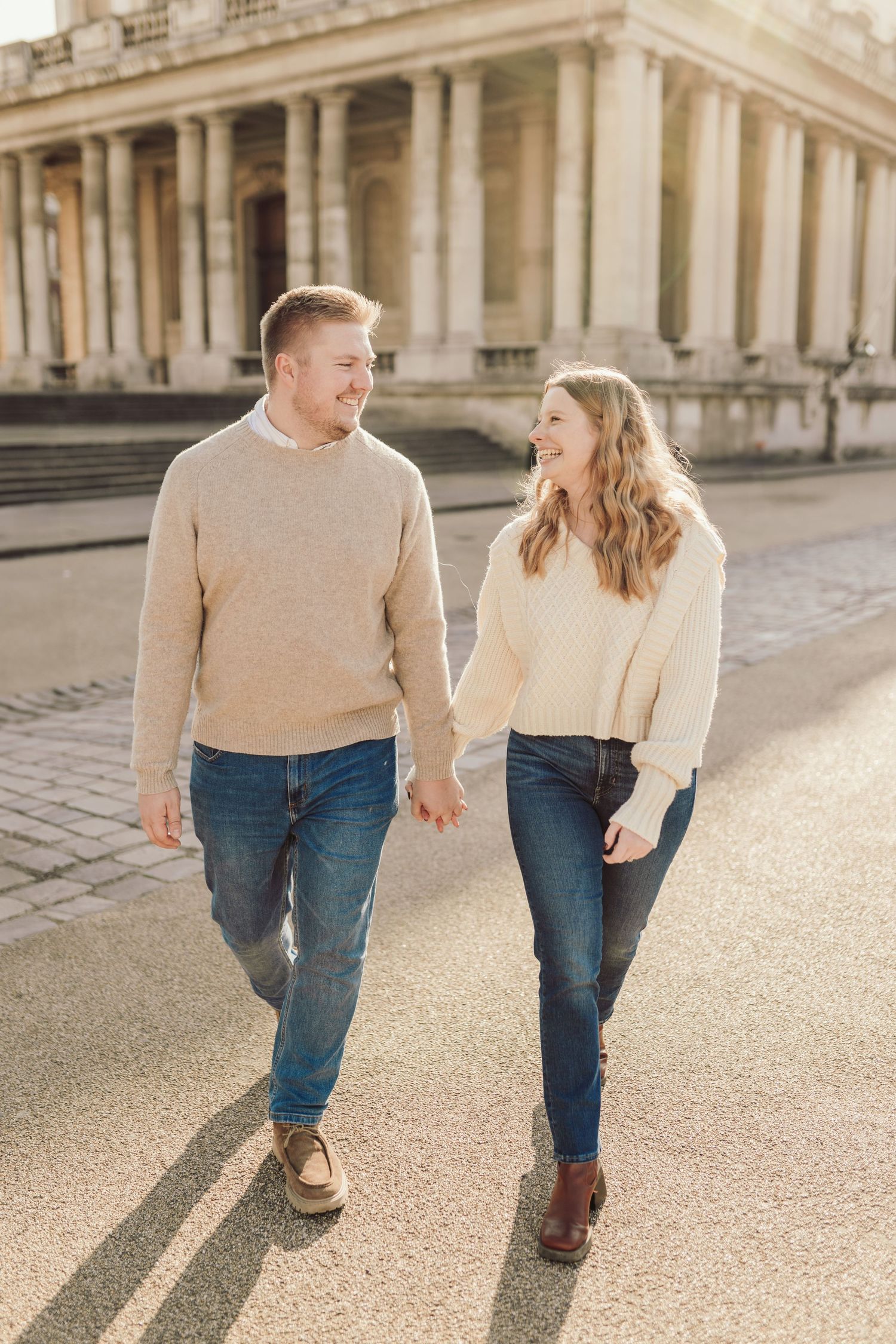 A couple in matching beige sweaters walks together outside a grand historic building.