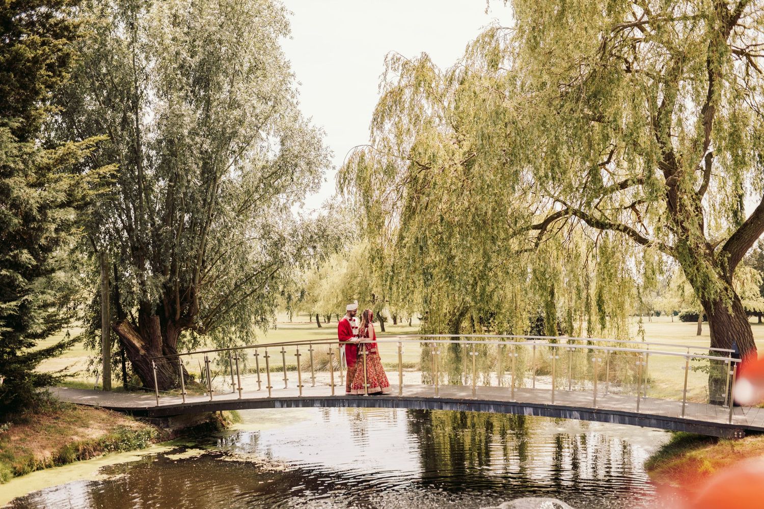A couple in traditional formal wear stands on an arched bridge over a pond surrounded by weeping willow trees in a park.