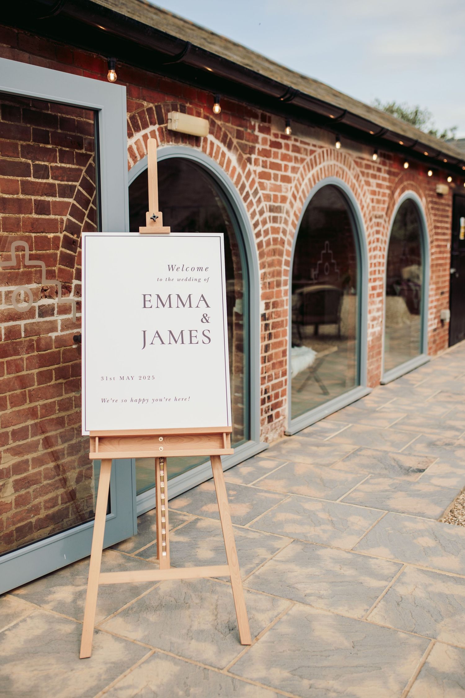 A wooden easel displays a welcome sign in front of a brick building with arched doorways and stone patio.