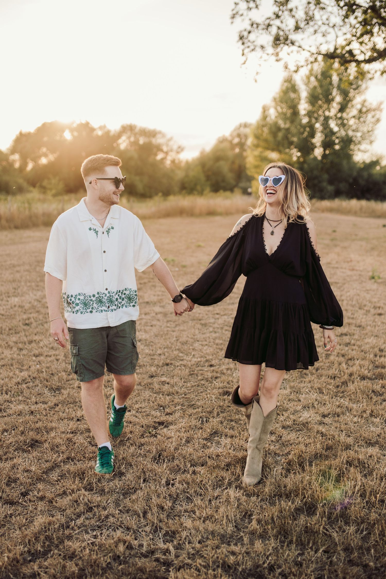 A stylish couple holds hands while walking through a sun-drenched field at golden hour.