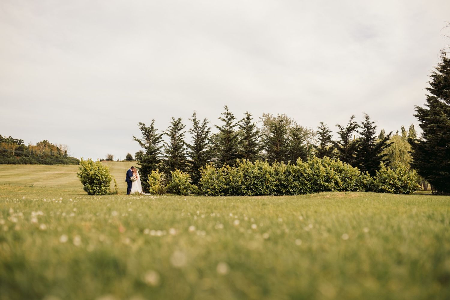 A couple walks among evergreen trees on a green hillside with overcast sky above the countryside landscape.
