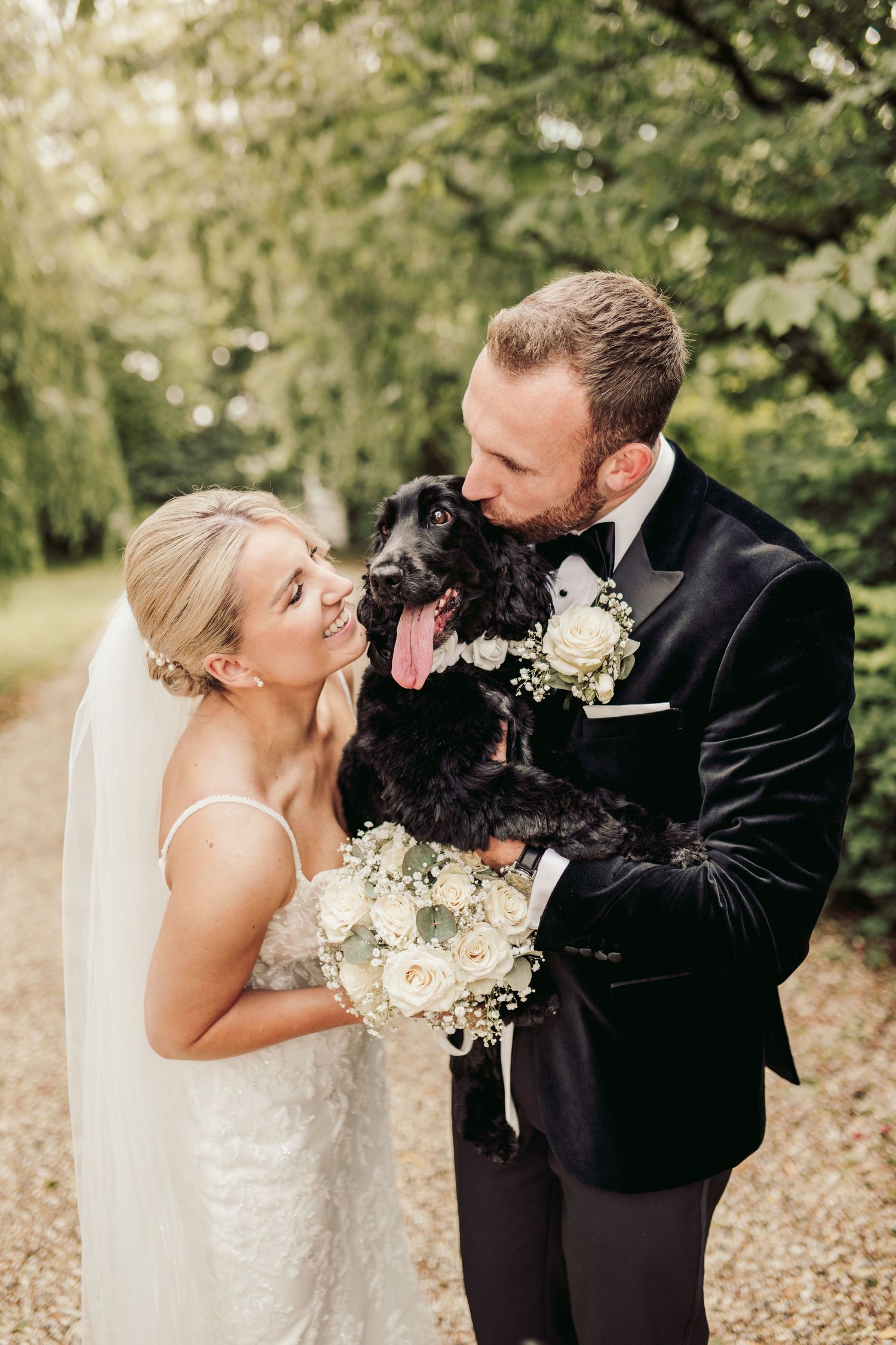 A happy couple in wedding attire shares a joyful moment with their black dog holding white flowers in an outdoor setting.