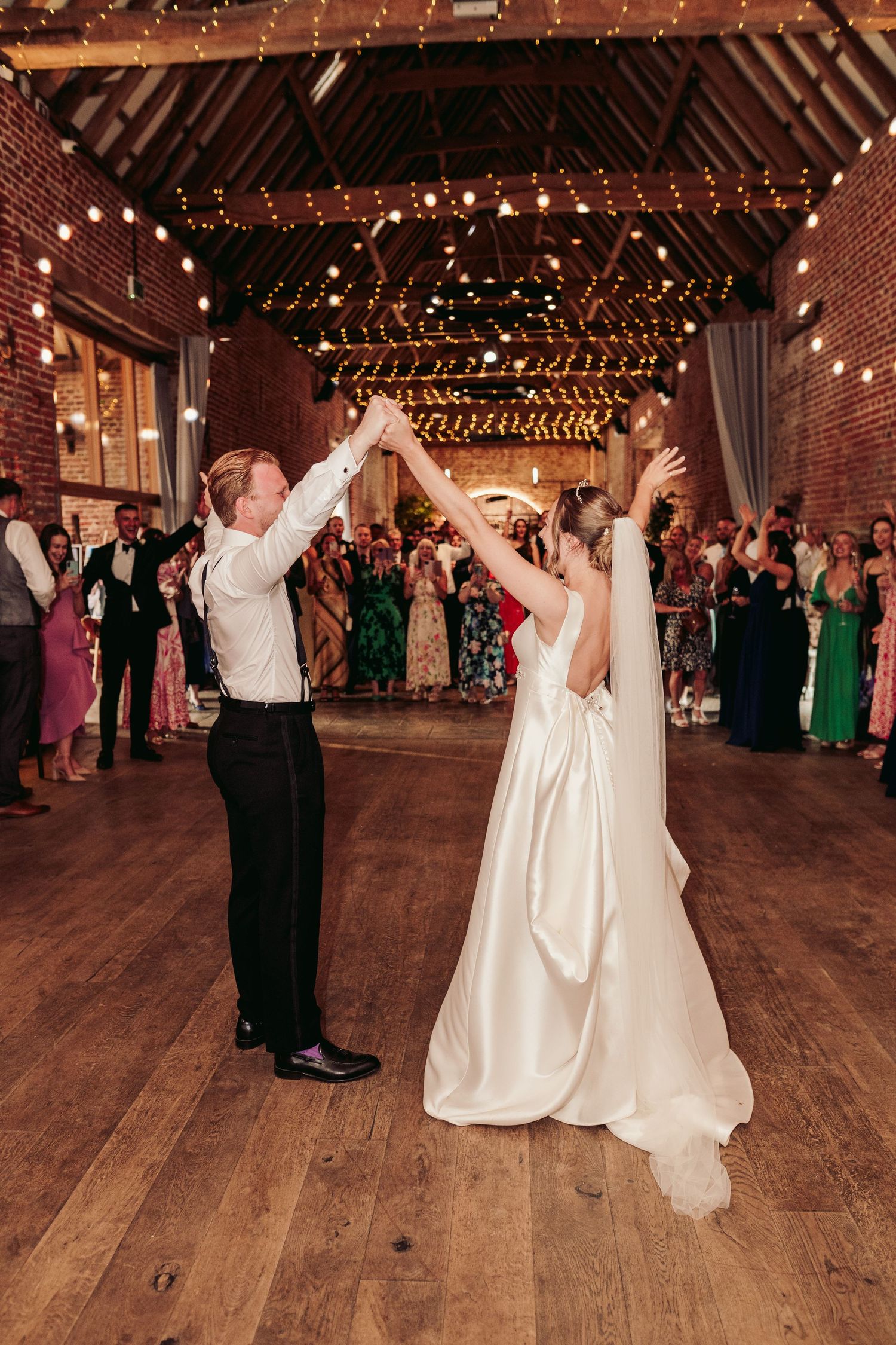 Couple dancing together at a rustic barn wedding reception with string lights overhead and guests watching.