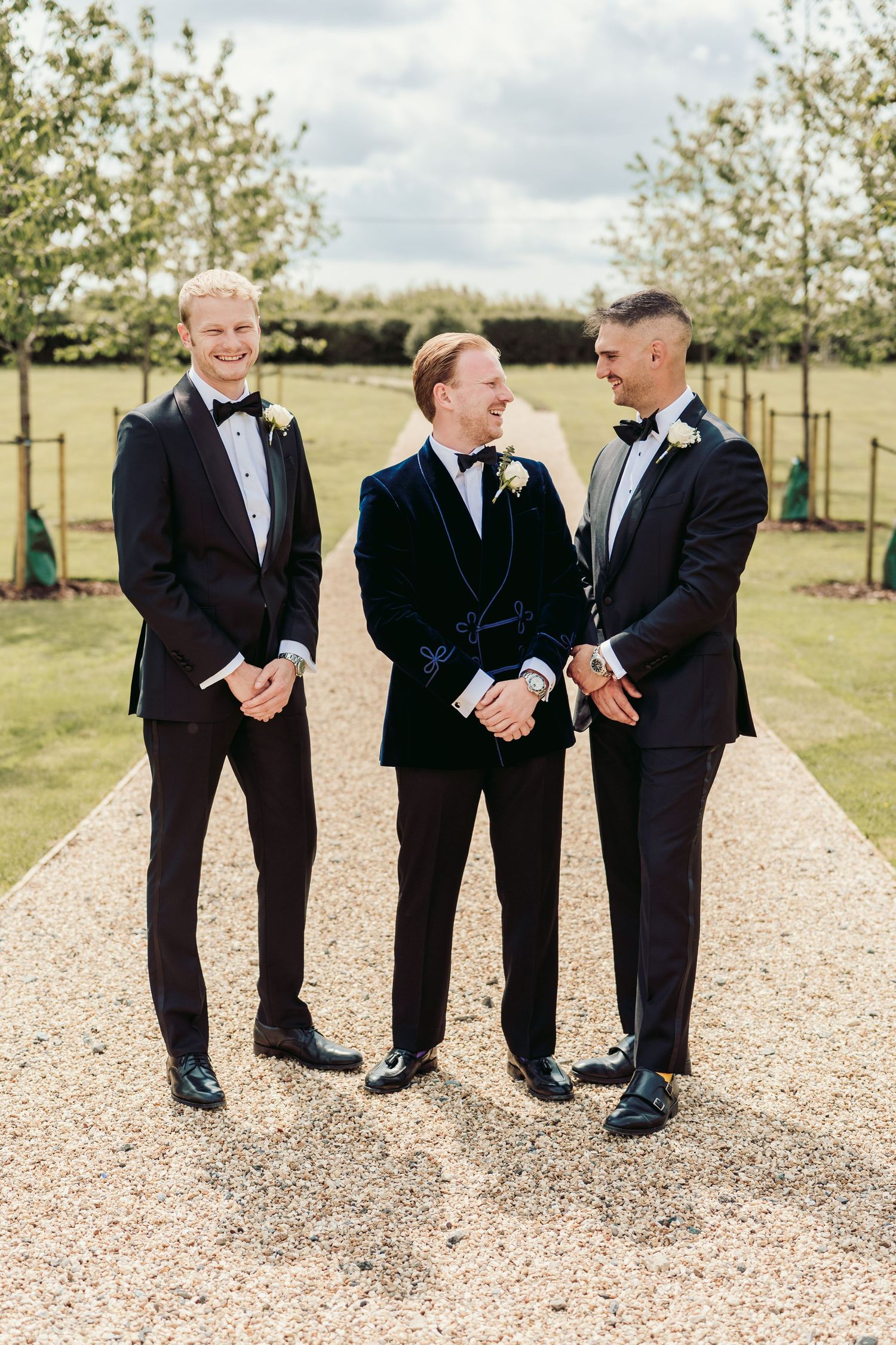 Three groomsmen in black tuxedos share candid laughs while standing on a gravel path during outdoor wedding photos.