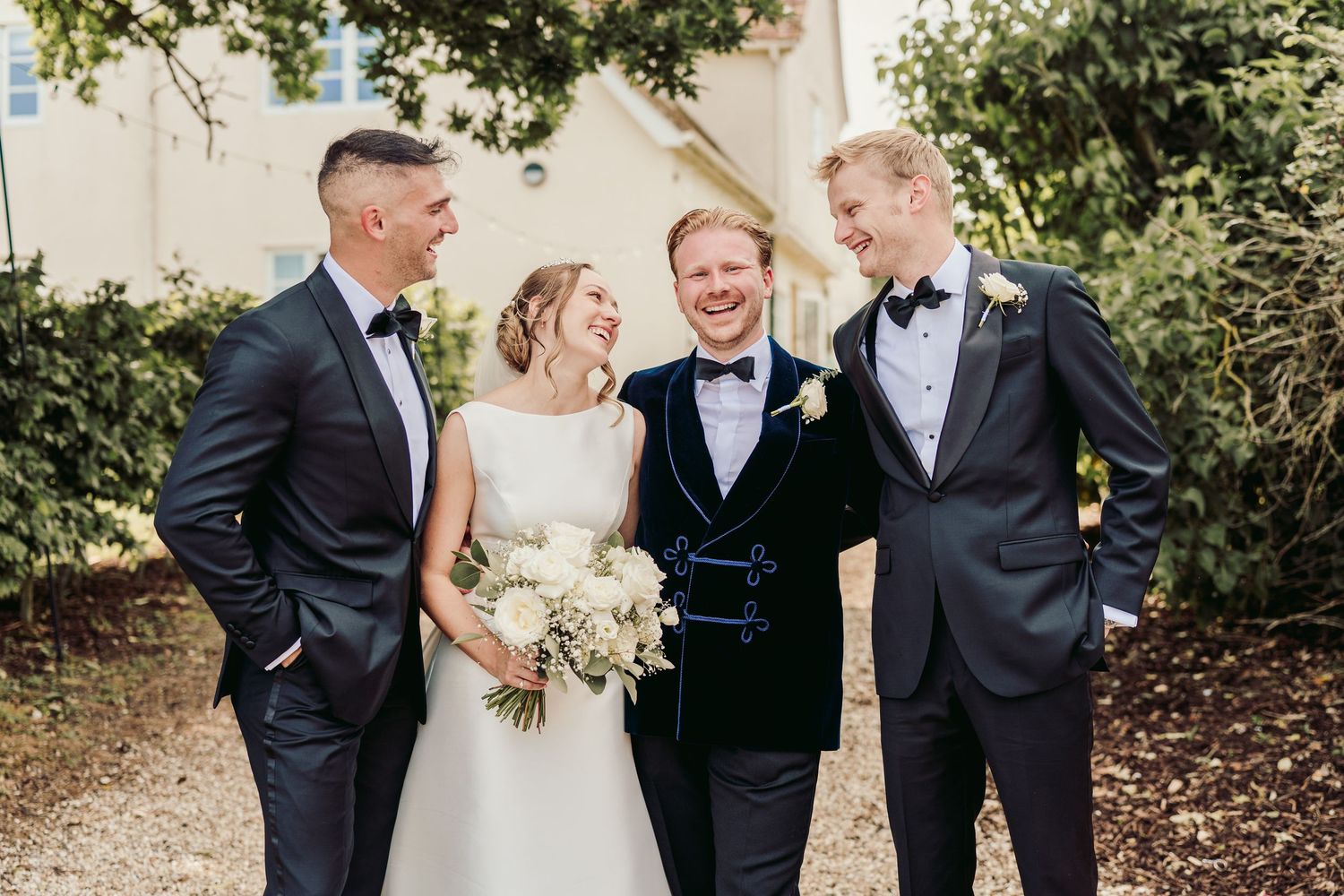 Wedding party members share a candid moment of laughter together outdoors under trees.