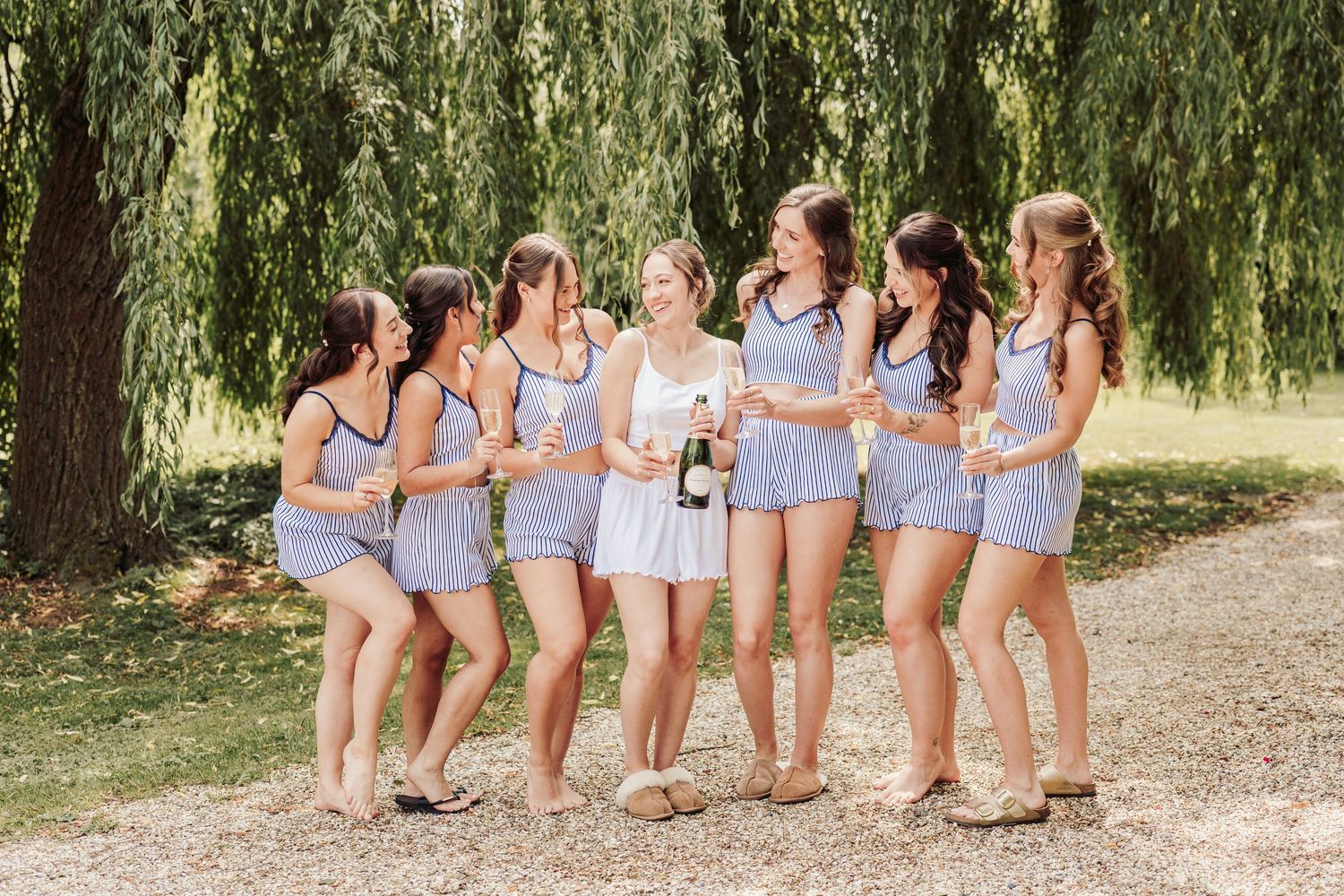 A group of bridesmaids in light blue dresses pose together on a path under willow trees.