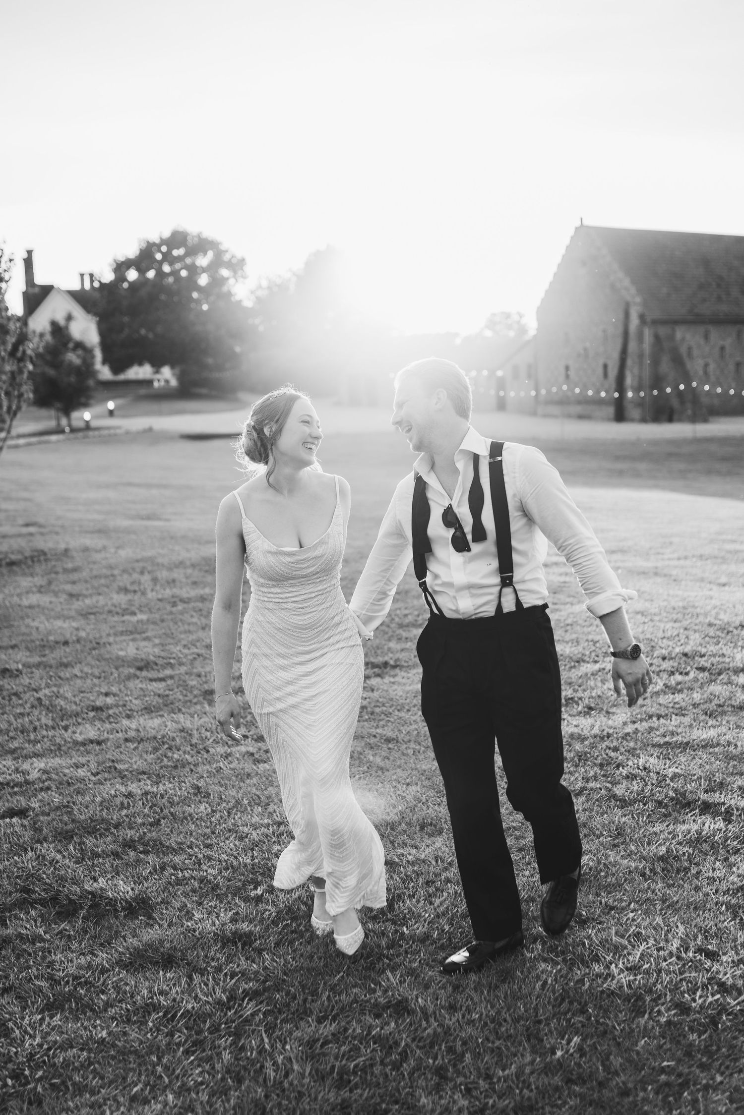 Black and white photo of newlyweds walking together on a sunlit grassy hill.