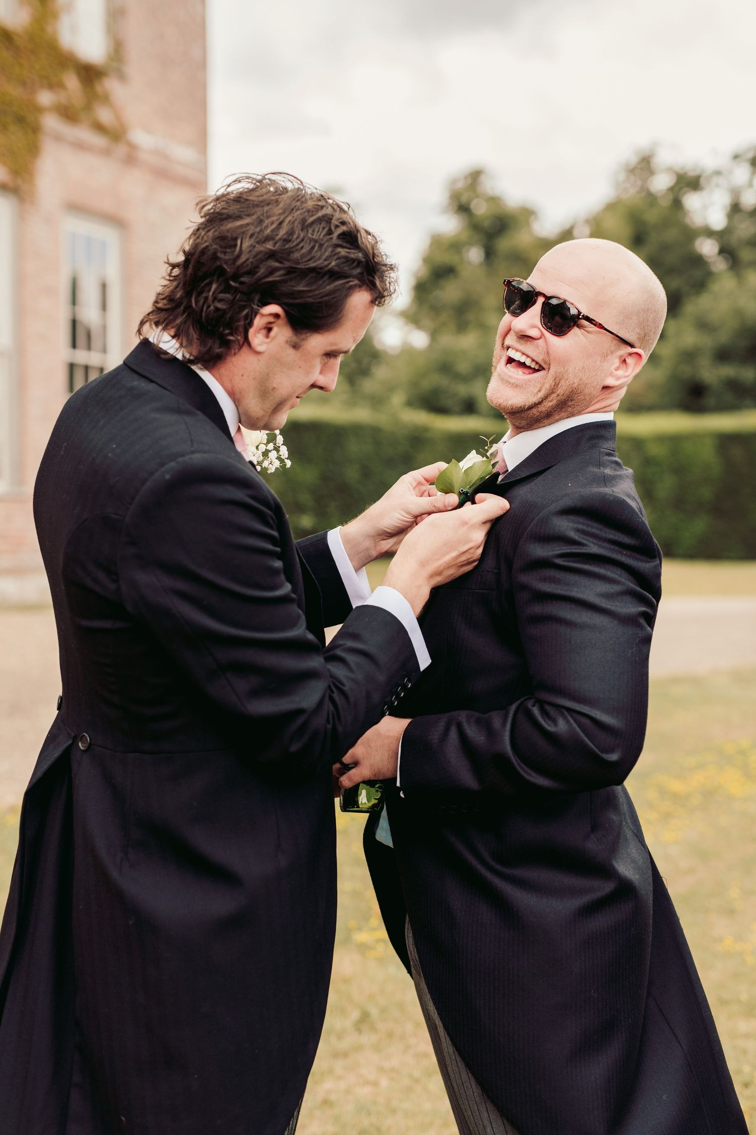 Two people in black suits share a joyful moment as one adjusts the other's boutonniere at an outdoor wedding.