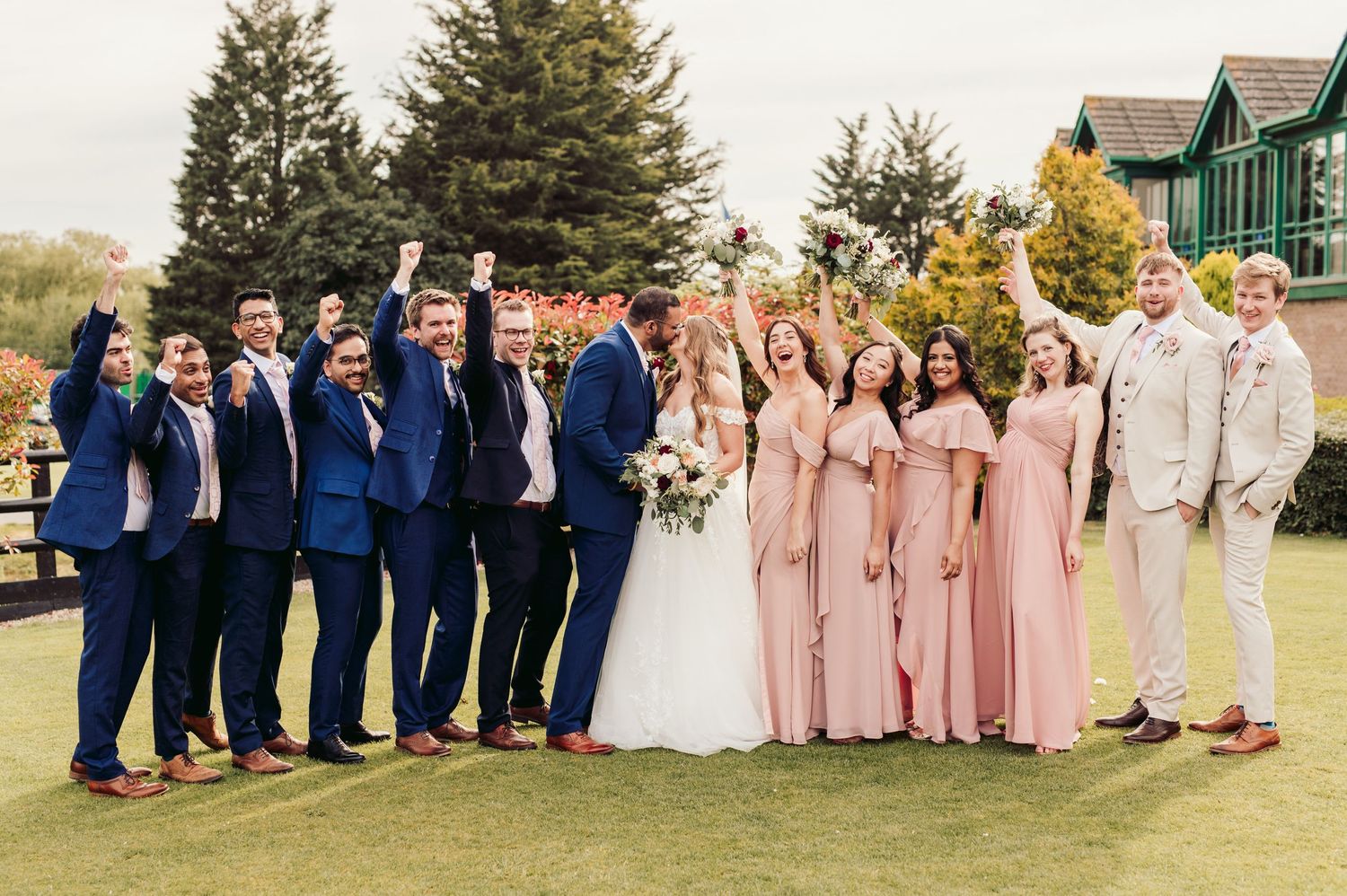 A wedding party poses outdoors on grass, with bridesmaids in pink dresses and groomsmen in navy suits.