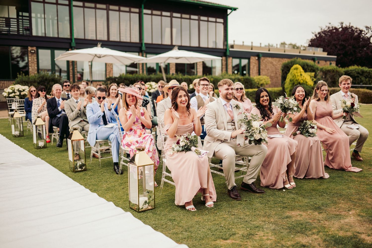 Wedding guests seated outdoors on white chairs watch ceremony on a grassy lawn with venue building in background.