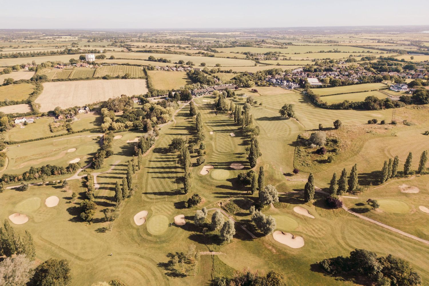 Aerial view of a golf course with green fairways, sand bunkers and trees scattered across rolling countryside terrain.