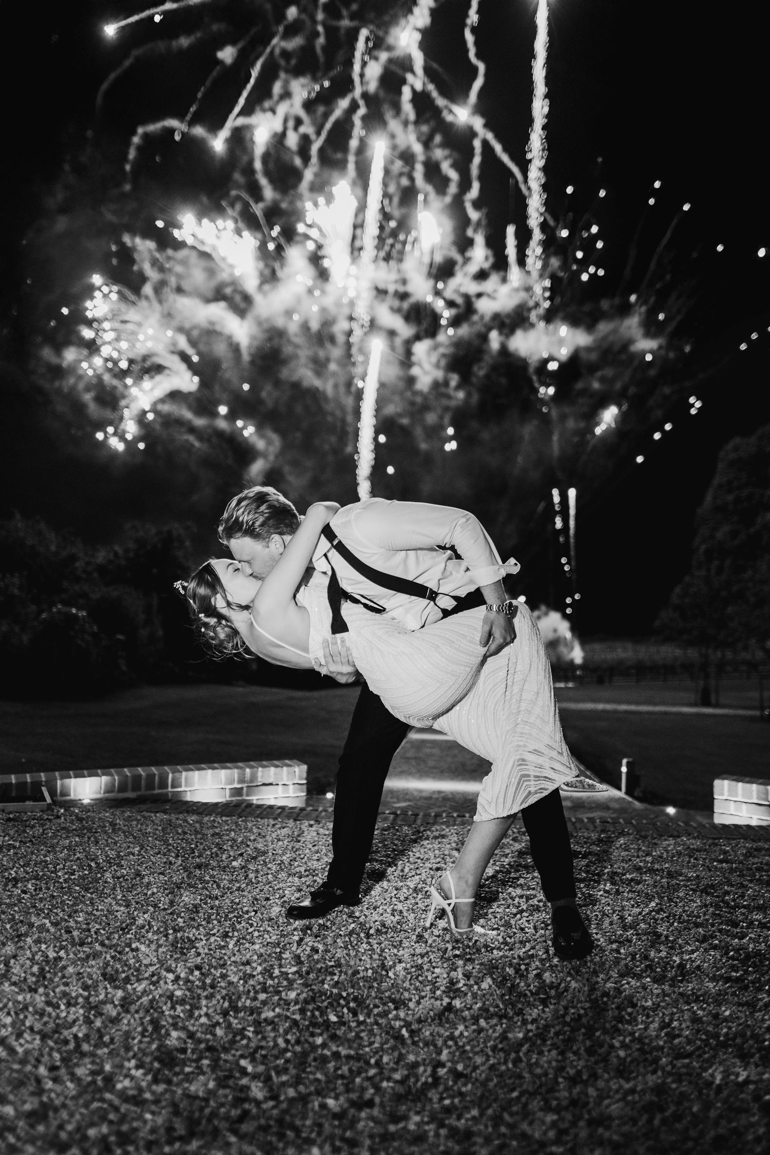 A couple performs a romantic dip pose against a backdrop of fireworks in the night sky in black and white photography.