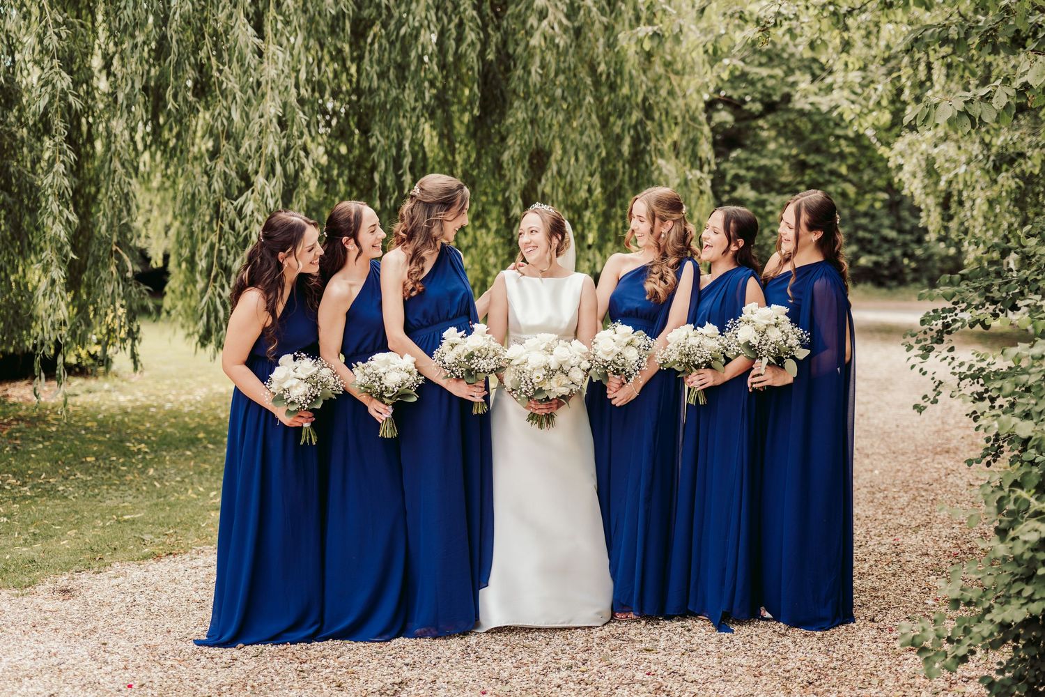 A wedding party in navy blue dresses and white gown poses together under weeping willow trees on a sunny day.