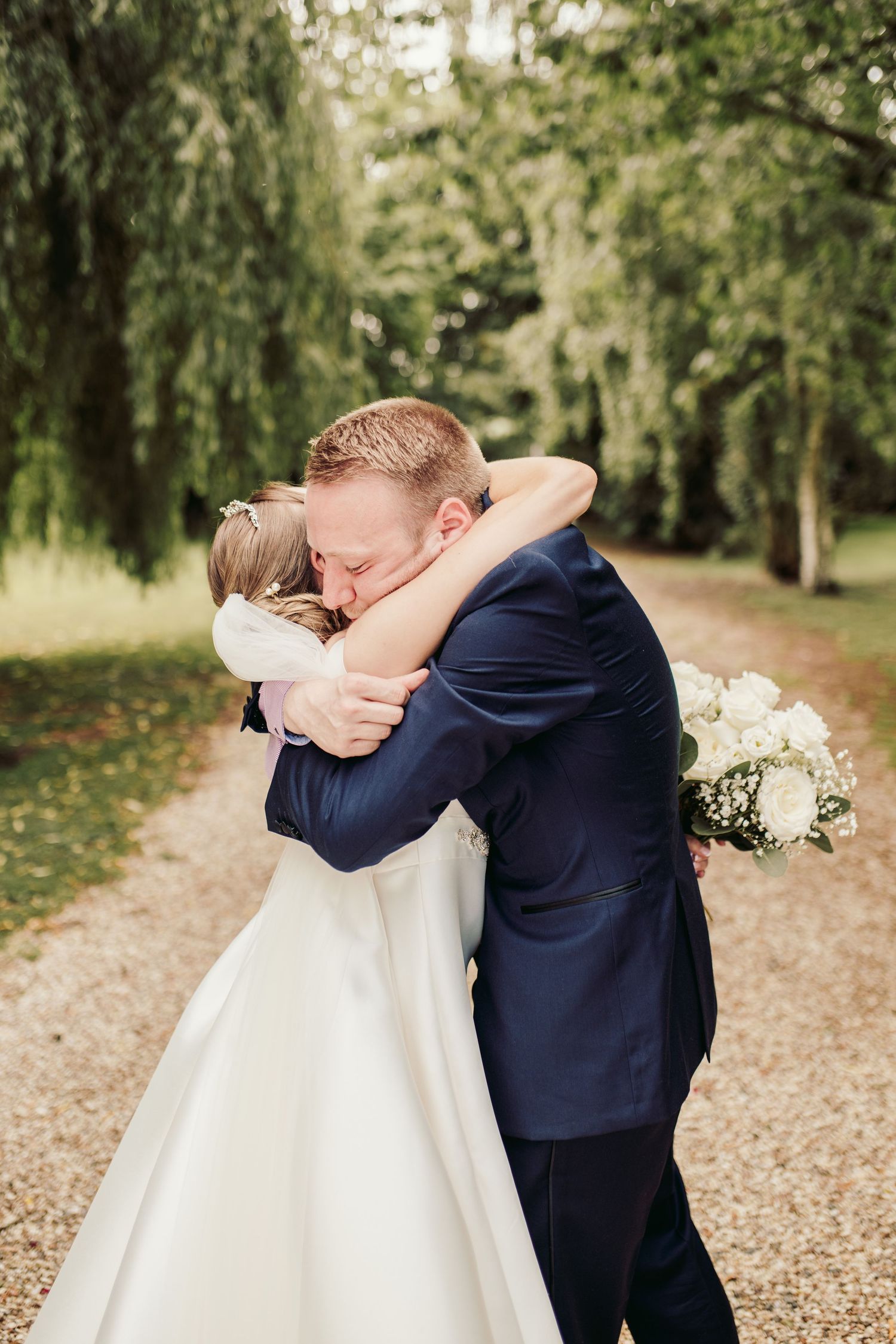 A couple in wedding attire embraces on a tree-lined path covered in fallen autumn leaves.