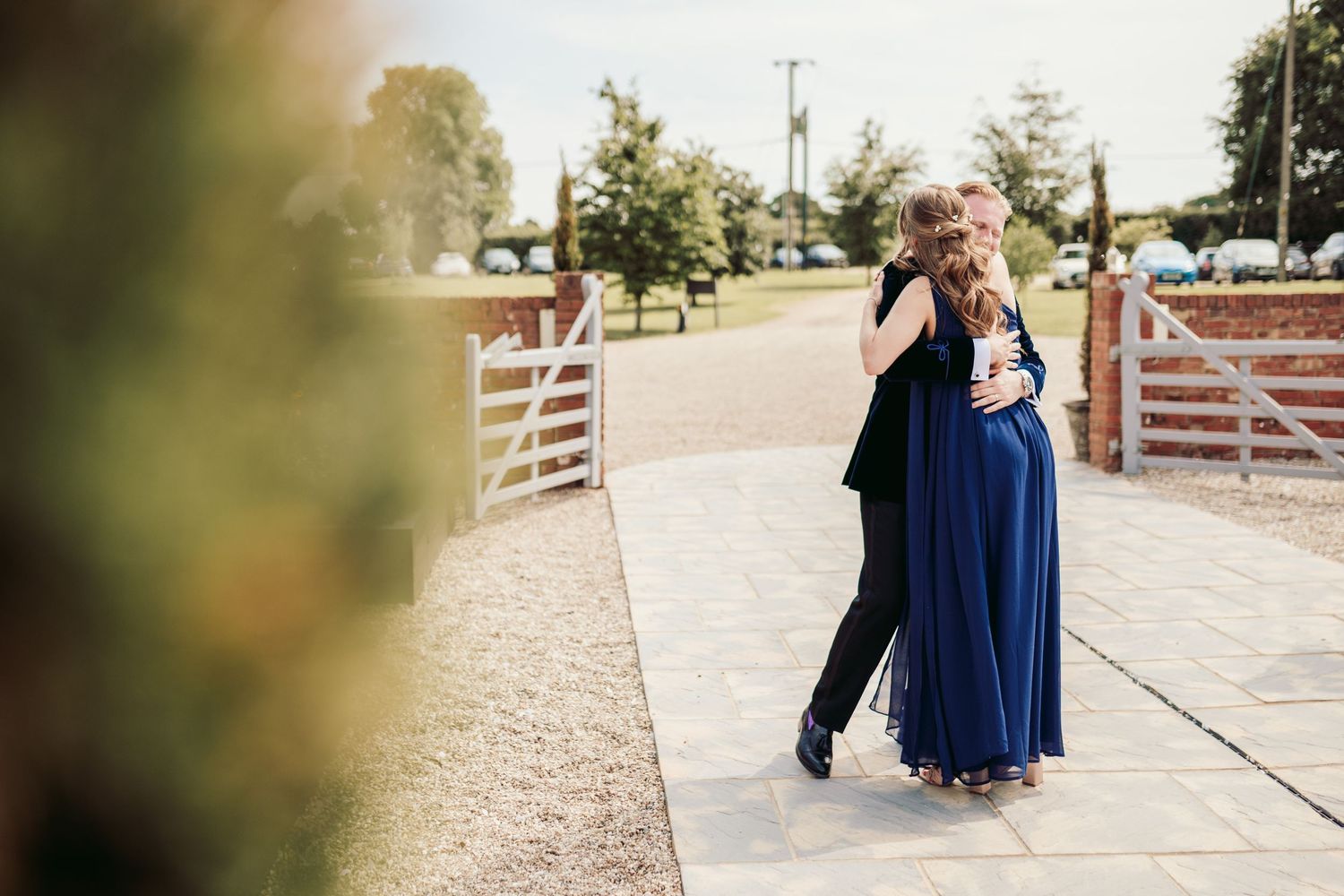 A couple in formal attire walks hand in hand down a gravel path at a rustic outdoor venue.