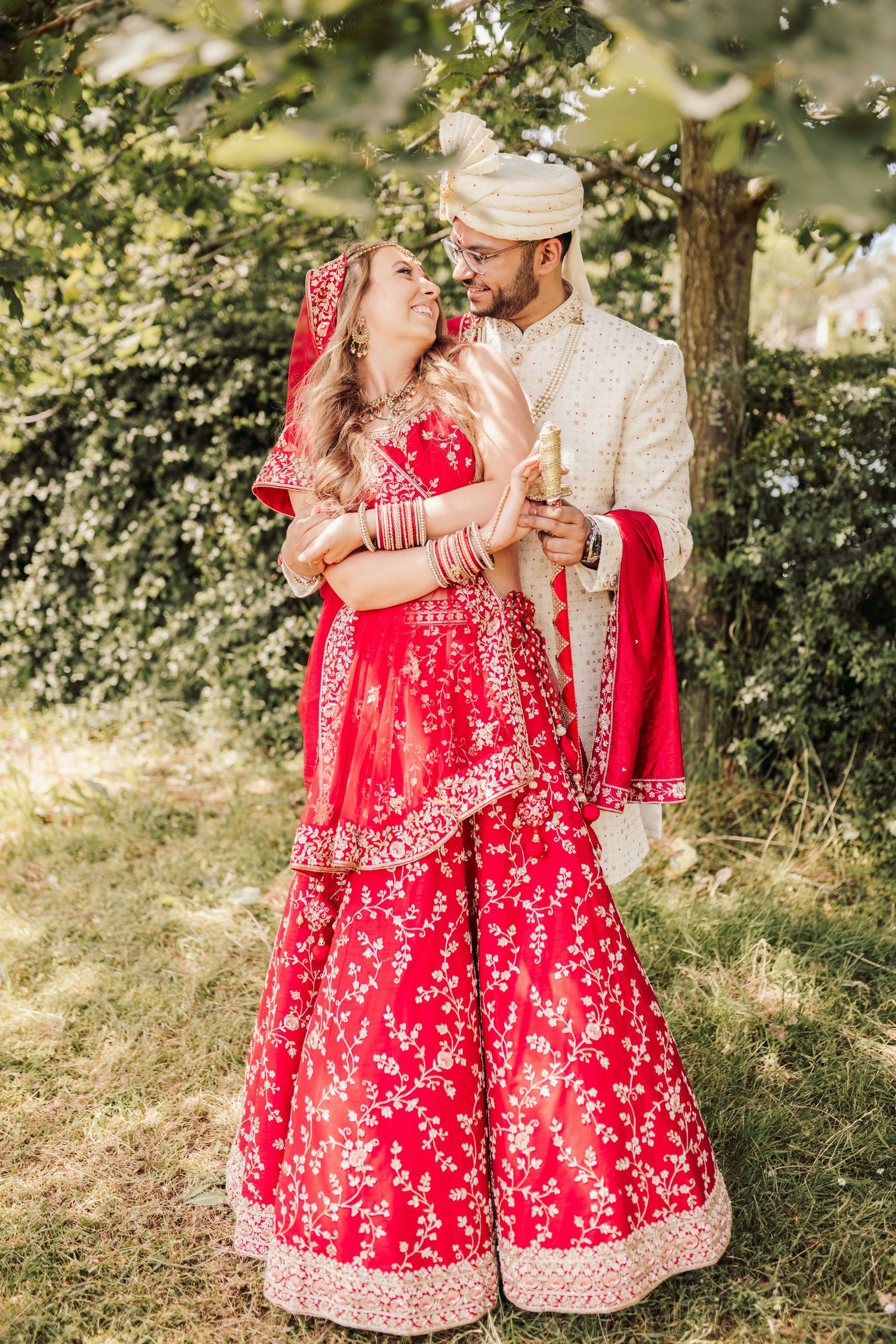 A couple in traditional South Asian wedding attire embrace outdoors, with the bride in a red embroidered lehenga.