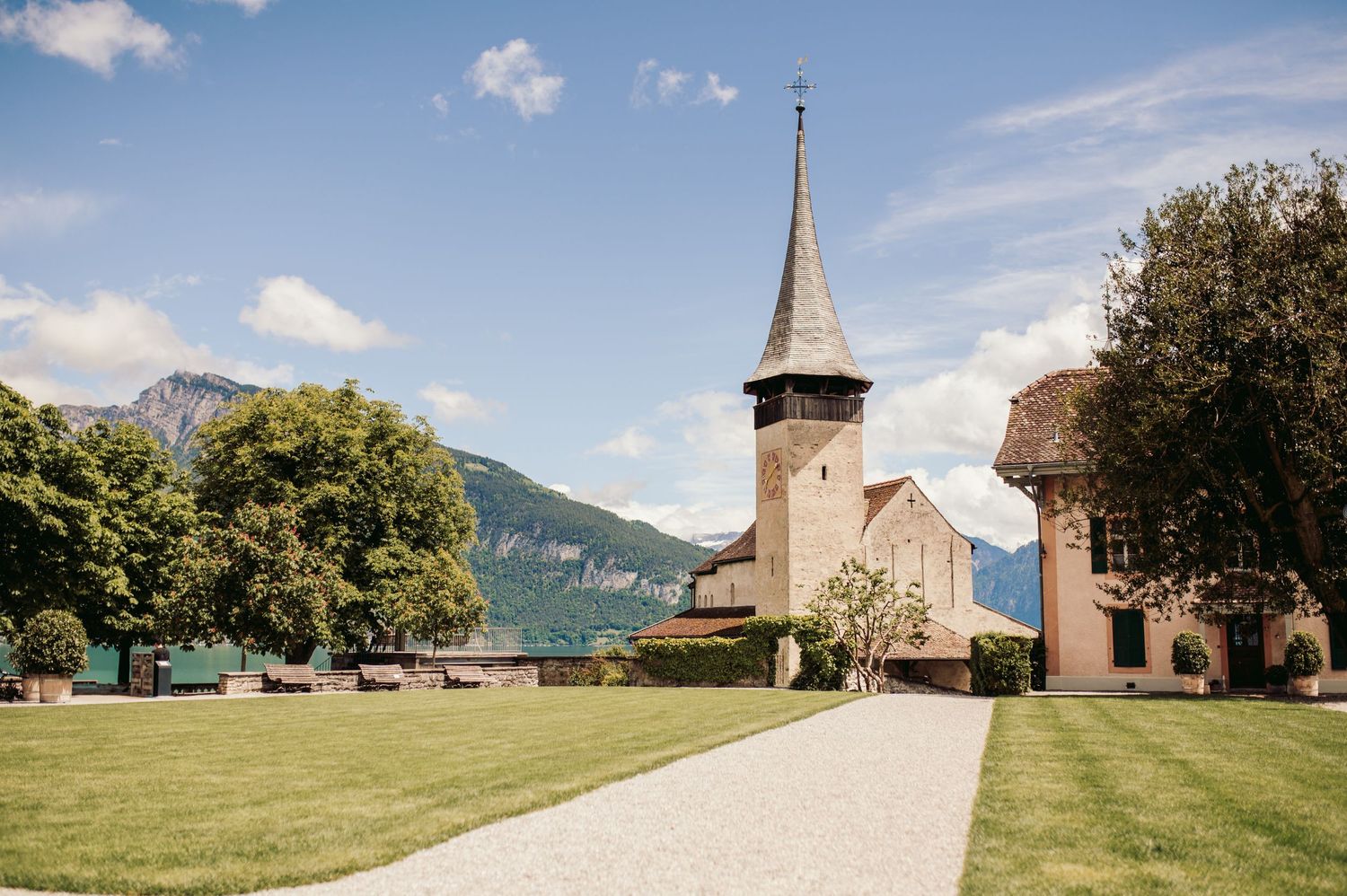 Church with tall spire along gravel path surrounded by green lawn and mountain landscape in background.Spiez switzerland destination wedding