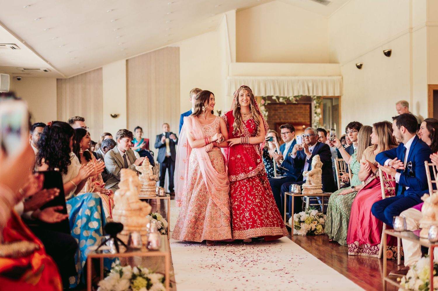 Two people in traditional Indian wedding attire dance together at a celebration while guests watch from their seats.