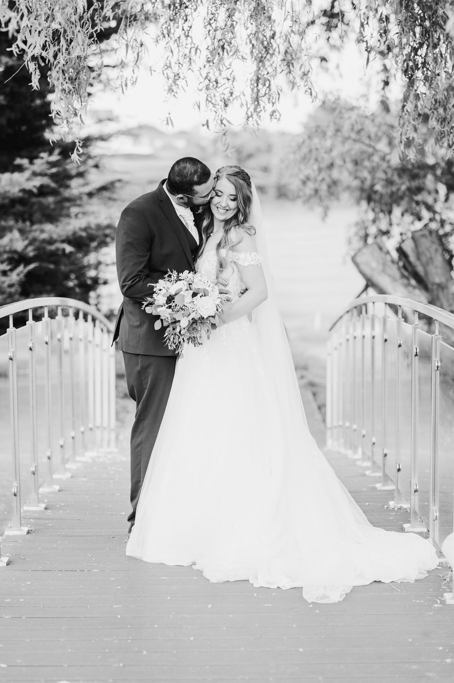 Black and white wedding photo of newlyweds embracing on a bridge with willow trees and floral bouquet.