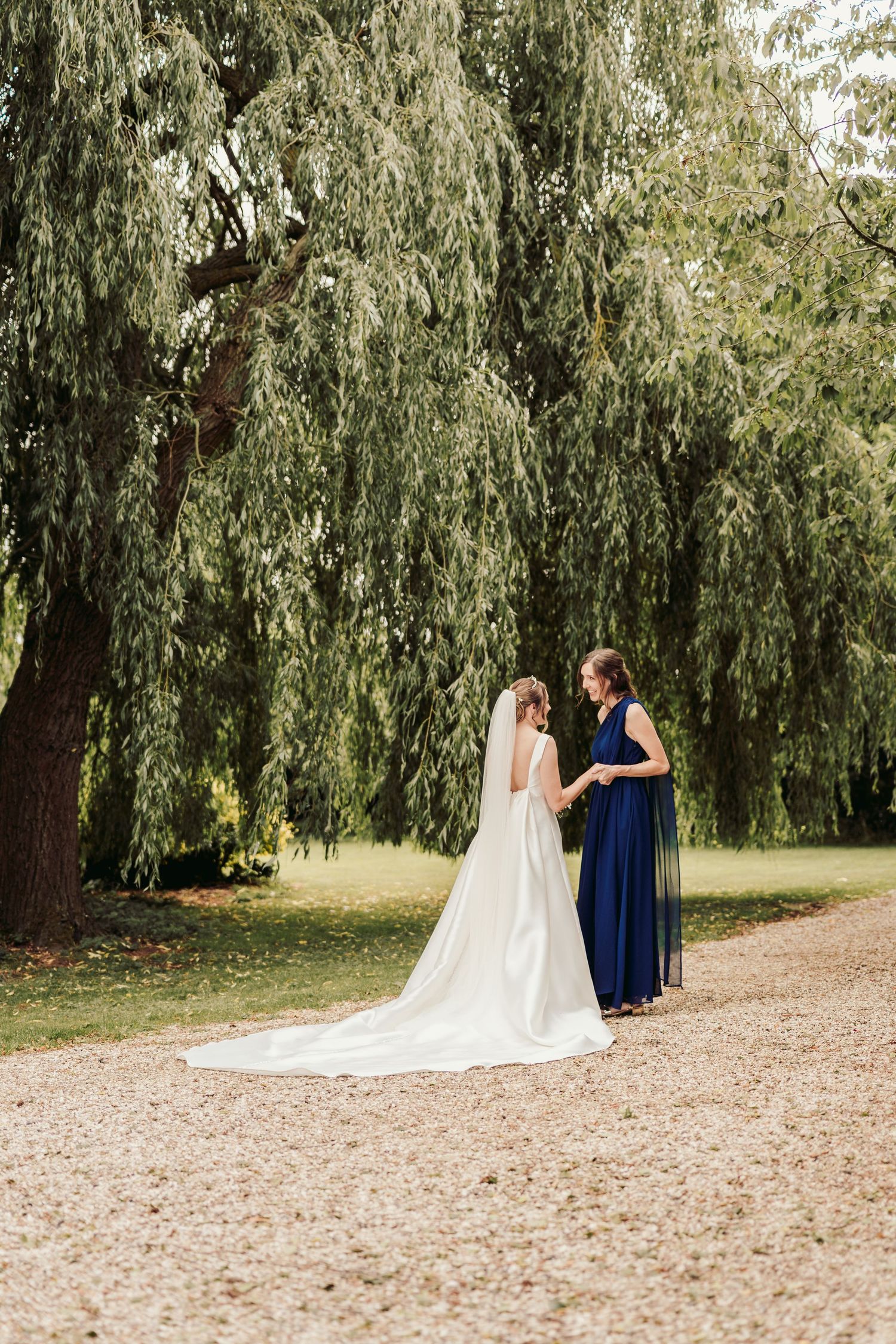 Two figures in elegant dresses stand together under a weeping willow tree on a gravel path during an outdoor event.